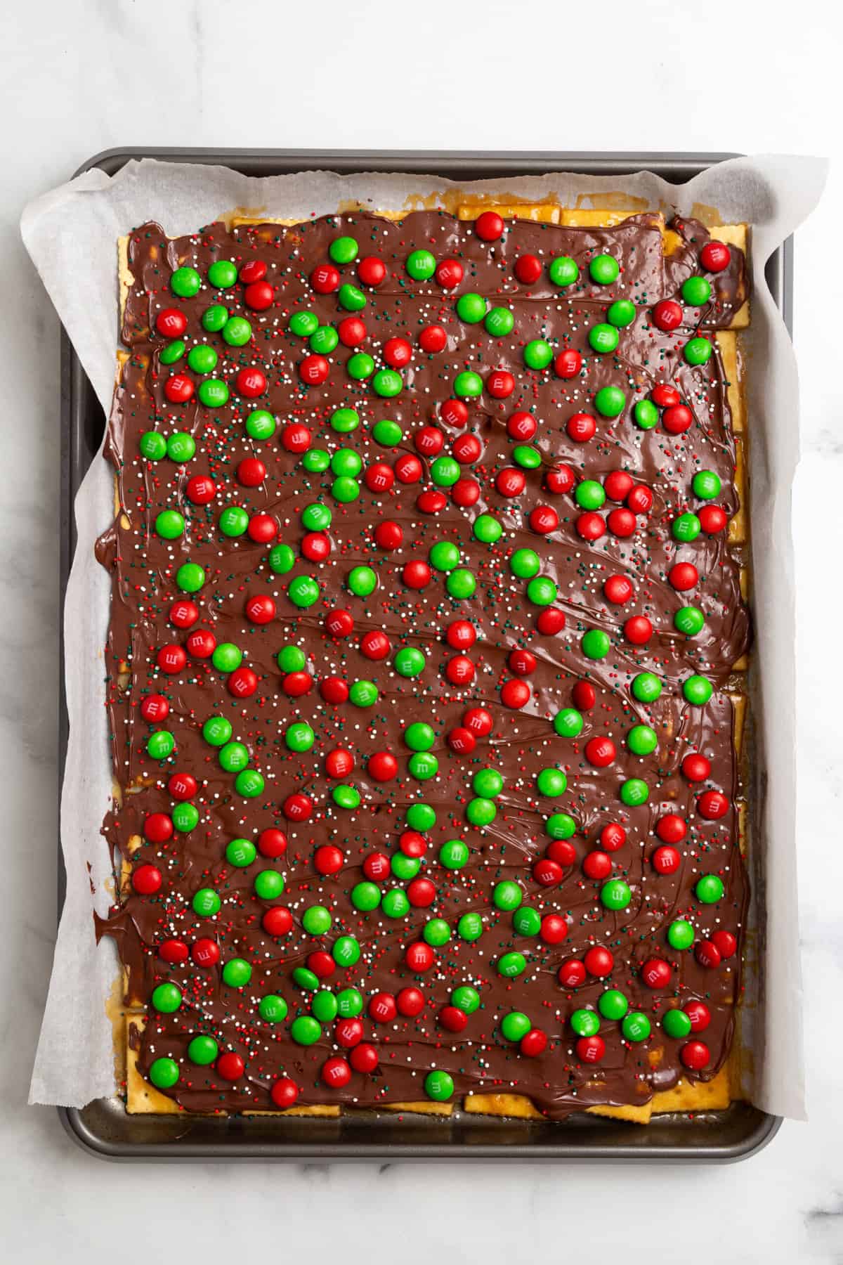 parchment-lined baking sheet with saltines arranged at the bottom, layered with caramel, melted chocolate, and green and red m&ms on top.