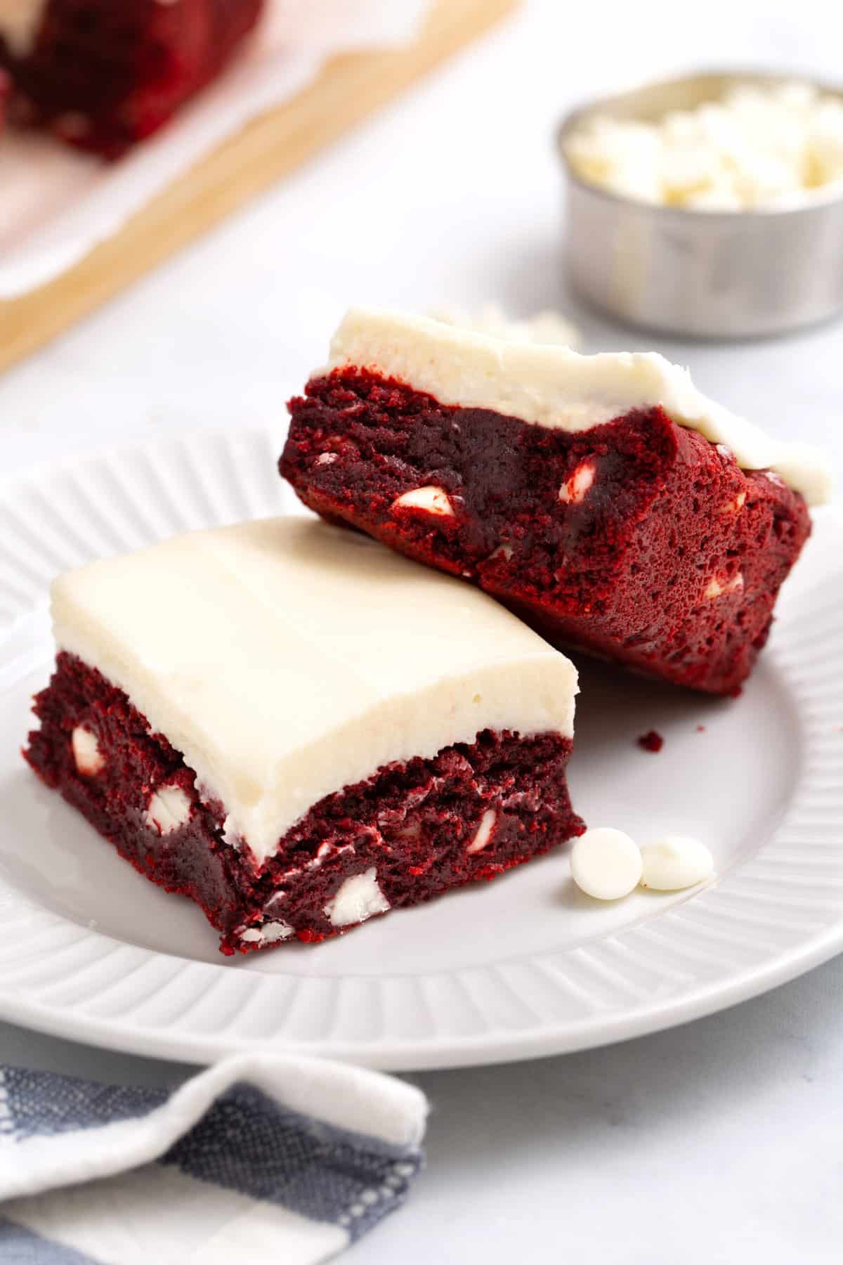 close up image of two square slices of dense red velvet brownies with white chocolate chips served on a white round plate. 