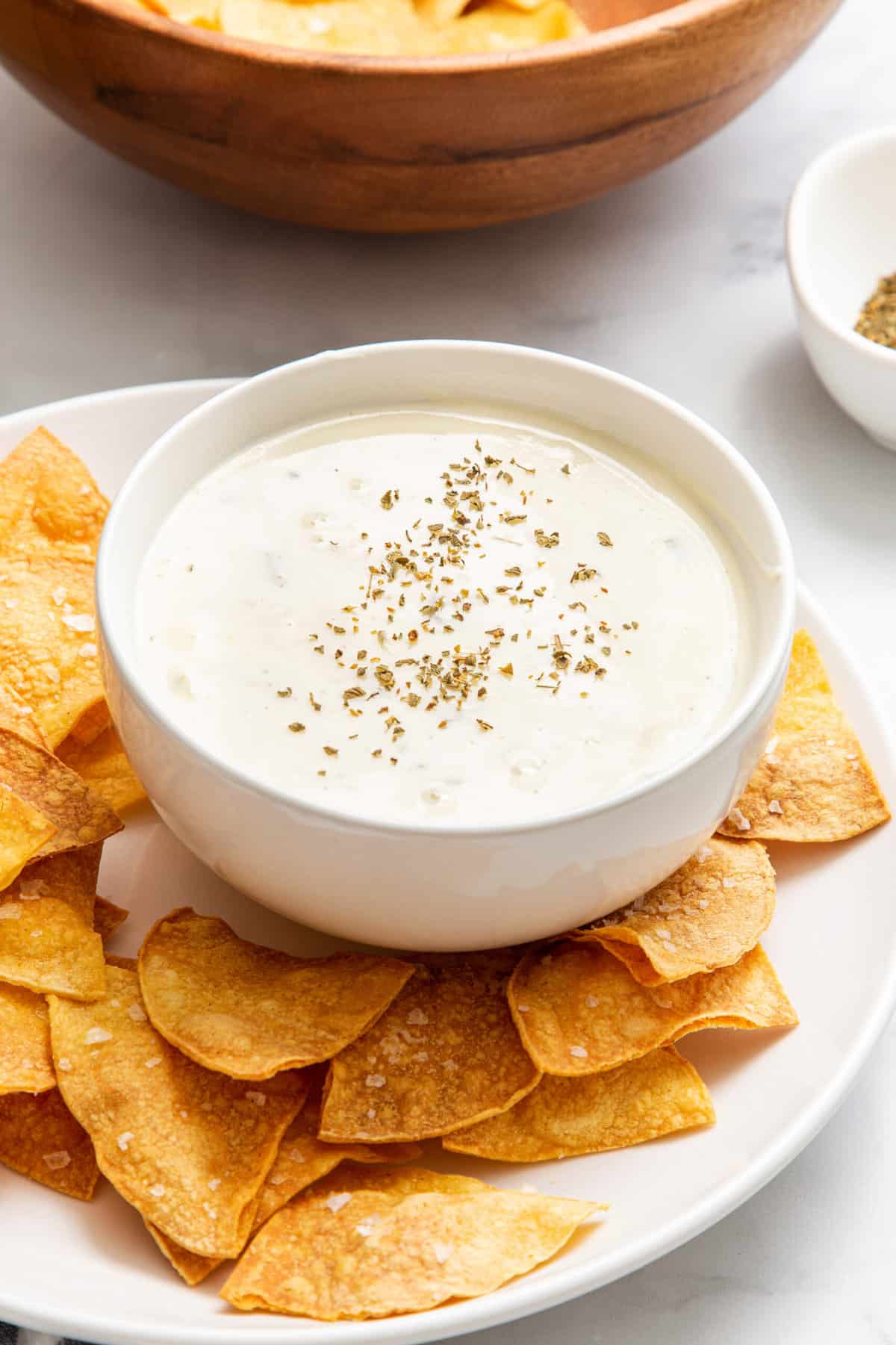 round white bowl with velvetta queso blanco sitting on a white round plate with homemade tortilla chips. 