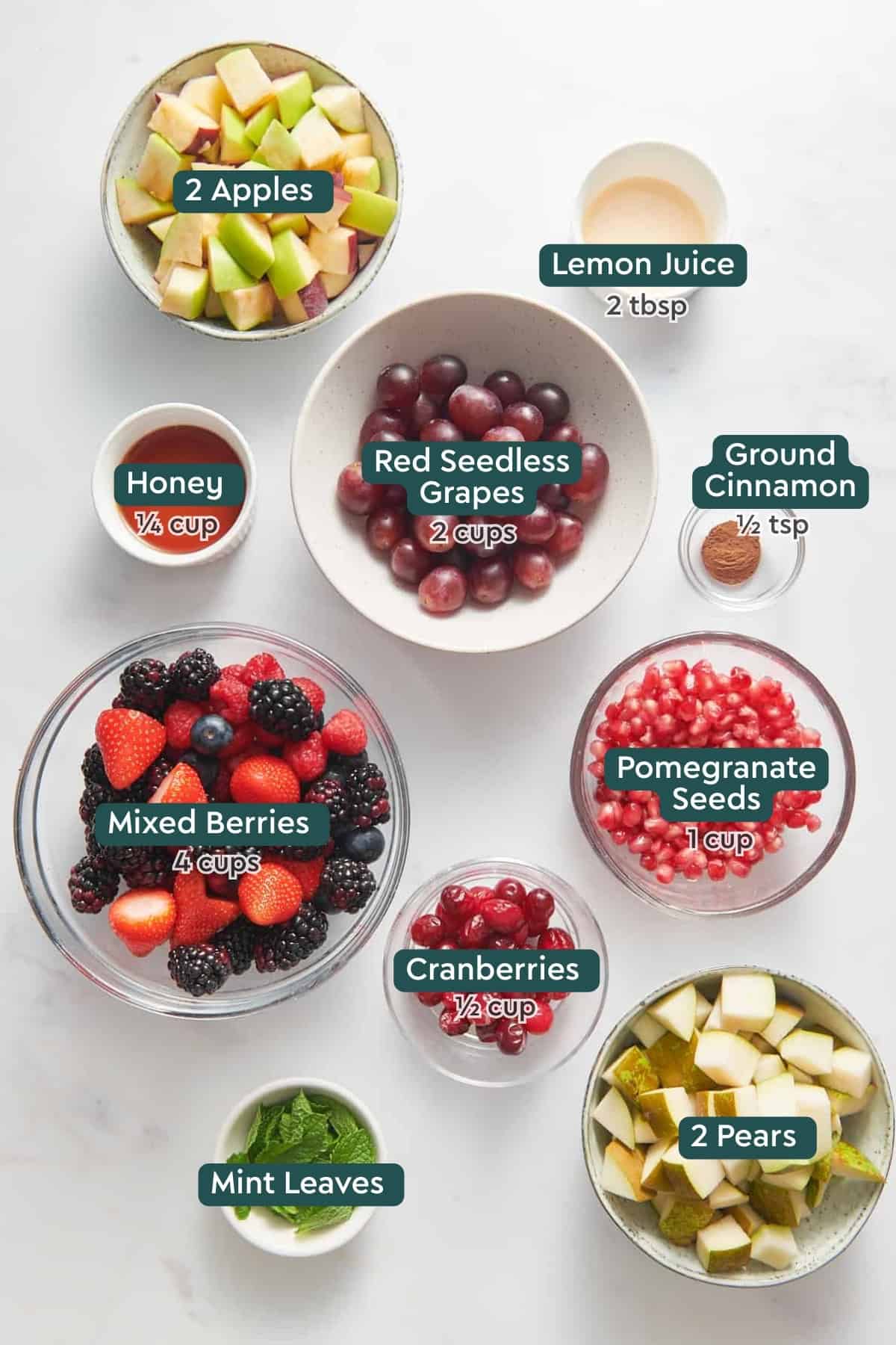 Ingredients for easy Christmas fruit salad, including apples, lemon juice, cinnamon, honey, grapes, pomegranate, berries, mint, and pears.