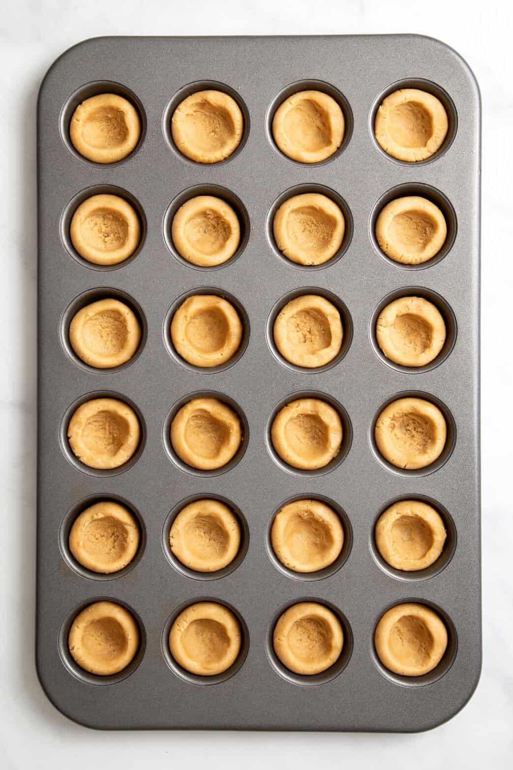 top down image of a 24-mini muffin tin with peanut butter cookie dough pressed in each spot.