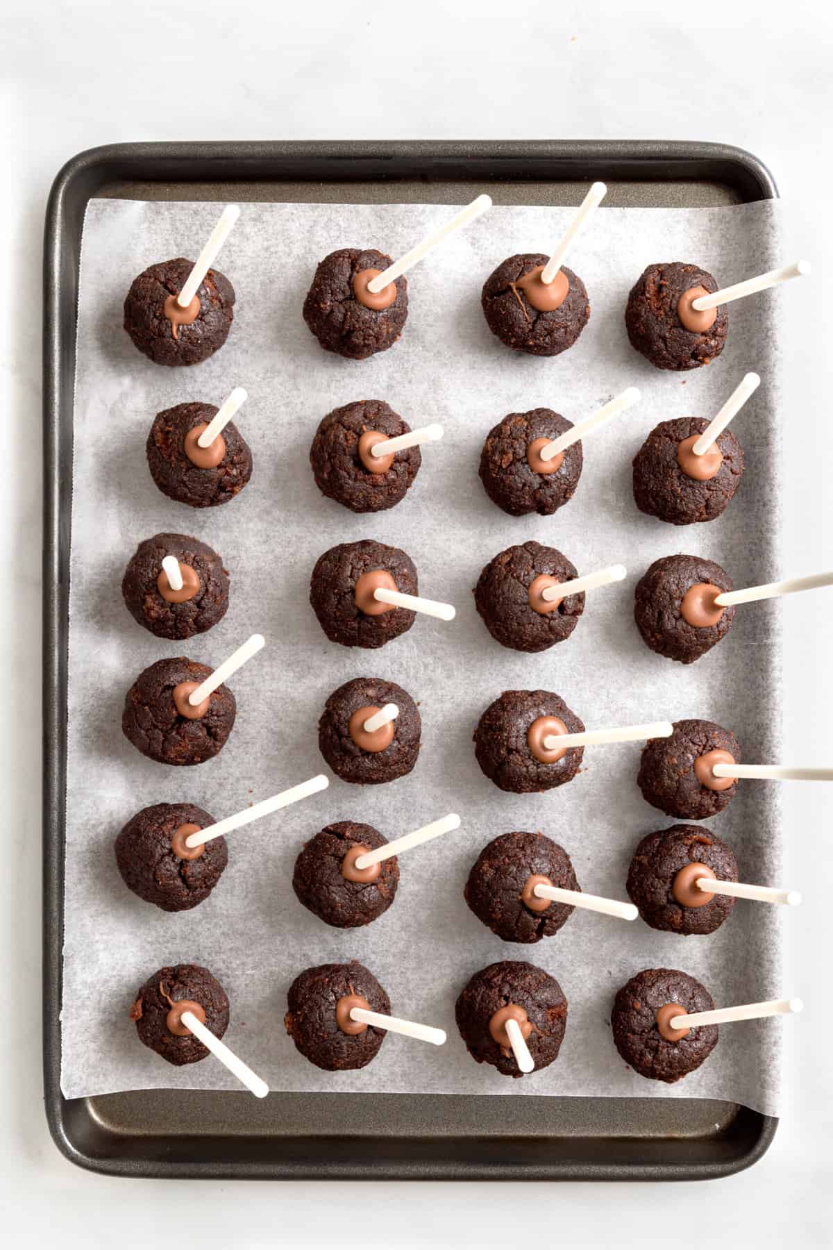 24 chocolate cake pops with sticks arranged on a parchment-lined baking tray. 