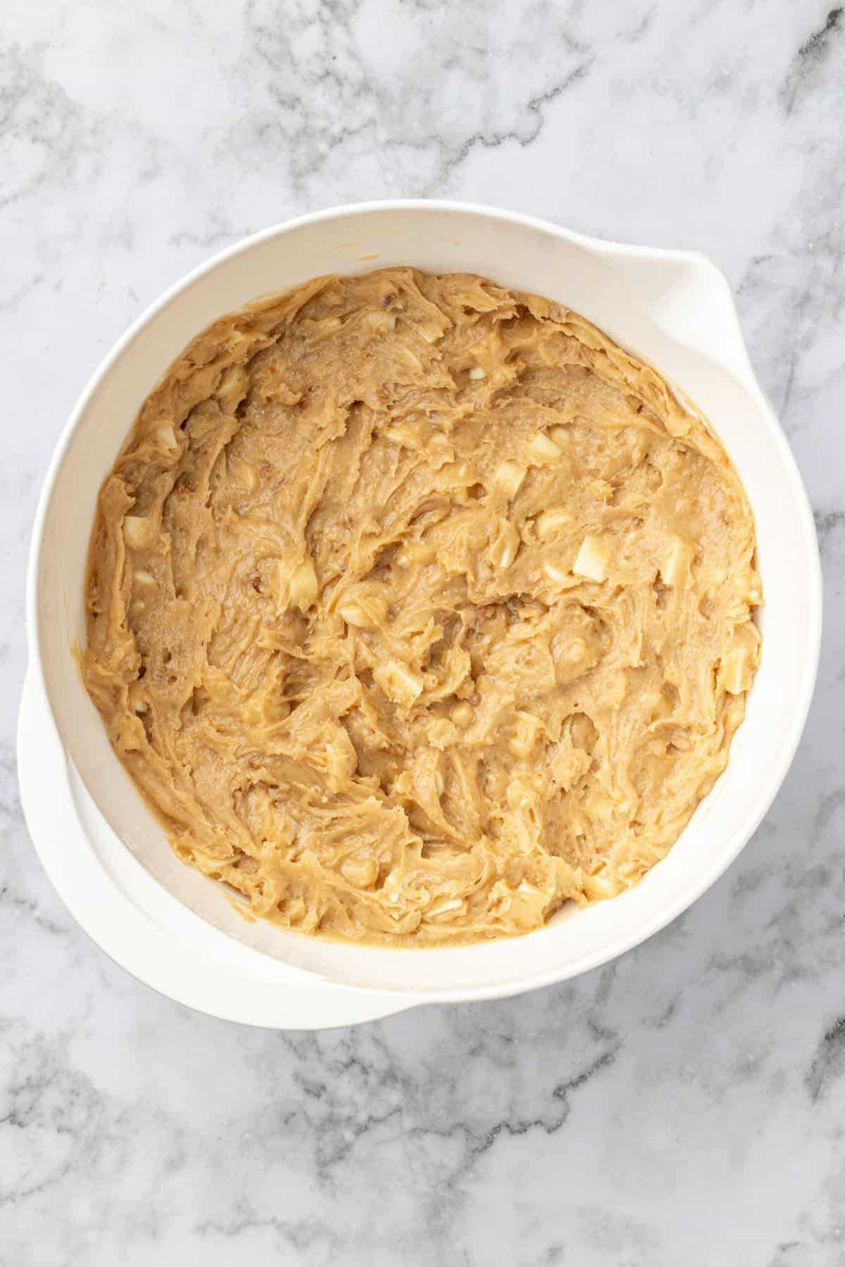 top down image of a white mixing bowl with combined flour, cornstarch, baking powder, salt, brown sugar, melted butter, eggs, walnuts, white chocolate chips and vanilla extract. 