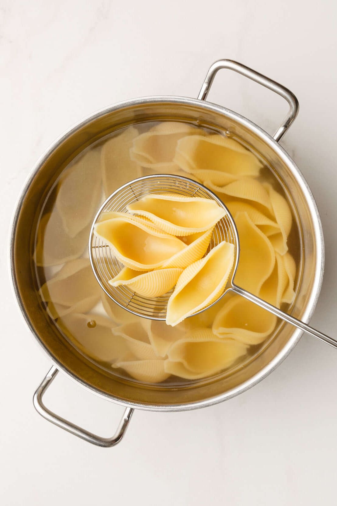 top down image of large pasta shells in a handheld strainer on top of a pot of pasta. 