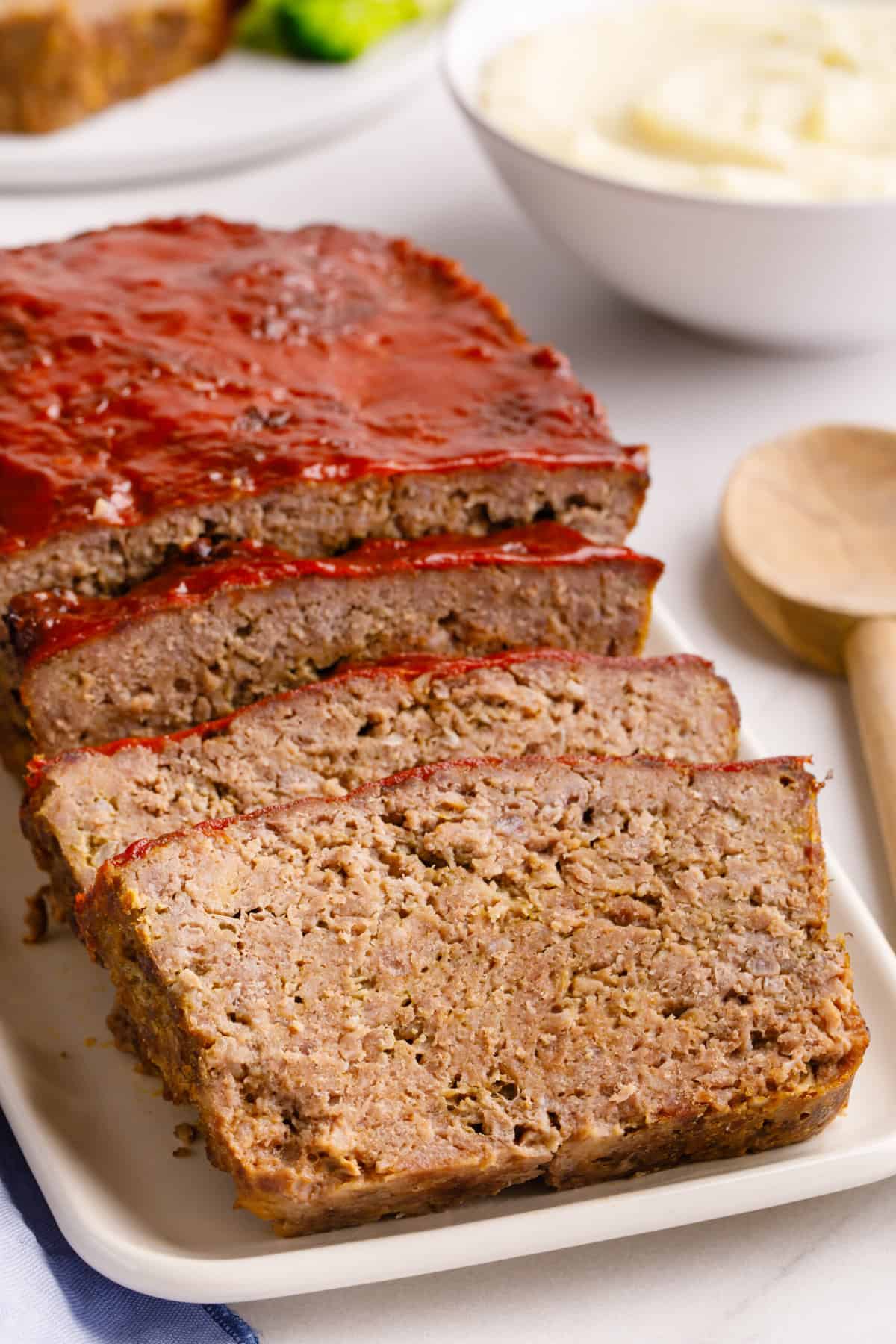 A slice of homemade Cracker Barrel meatloaf served on a white plate with mashed potatoes and broccoli in the background