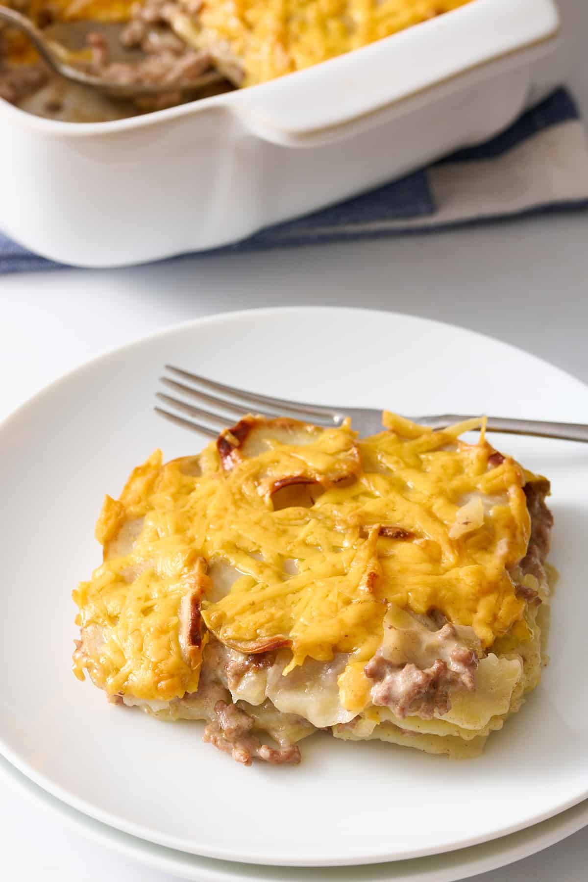 square serving of hamburger and potato casserole sitting on a white round plate with a silver fork. 