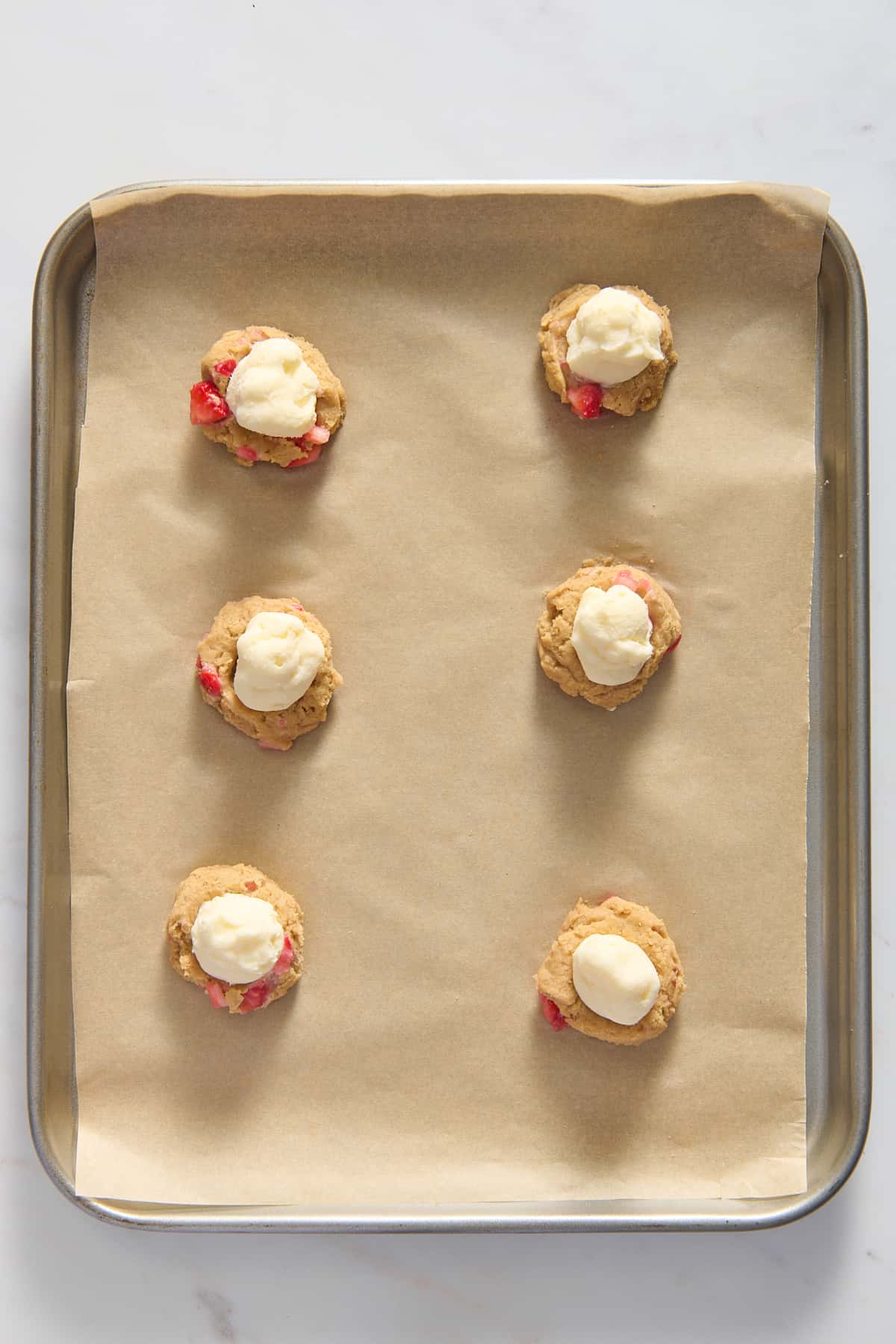 top down image of six strawberry cheesecake cookie dough balls with cream cheese filling prepared on a parchment-lined baking tray. 