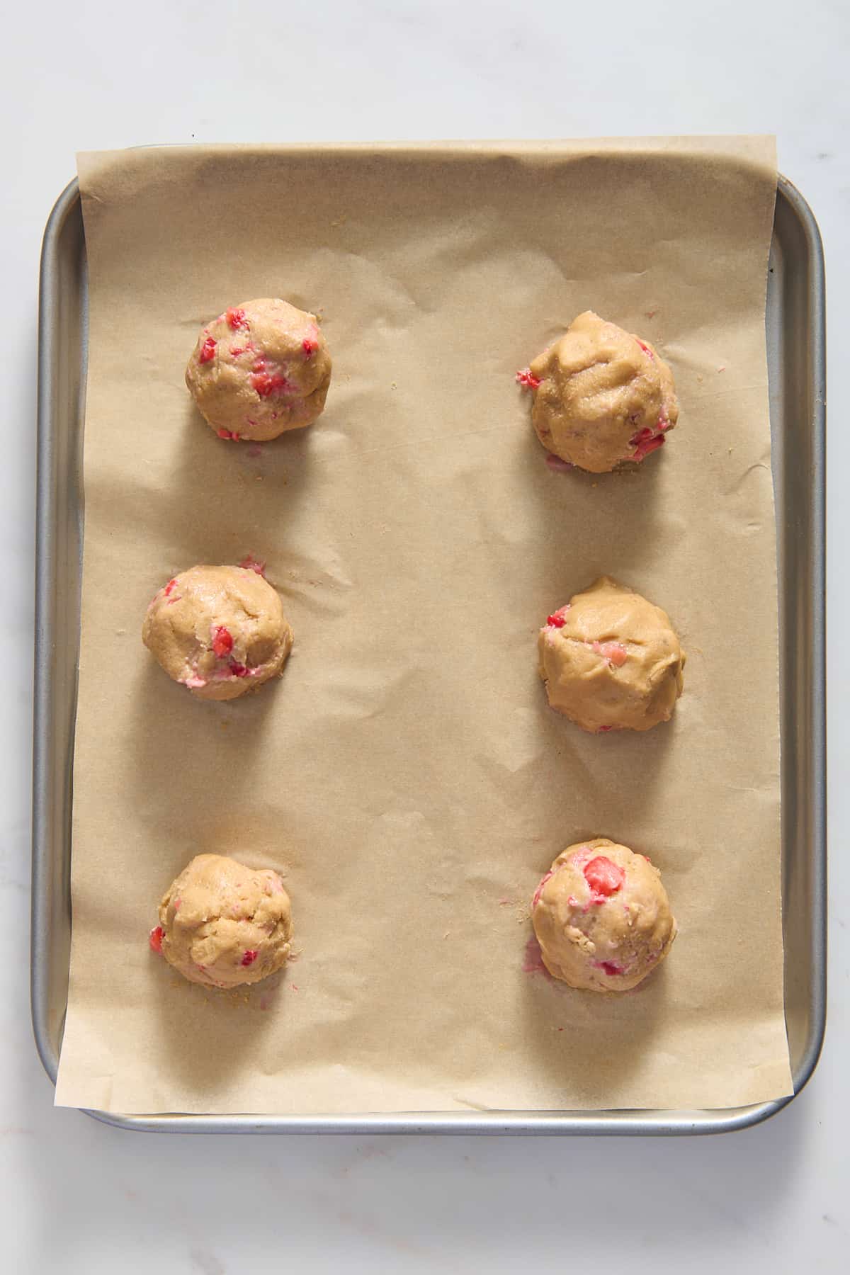 top down image of six strawberry cheesecake cookie dough balls prepared on a parchment-lined baking tray. 