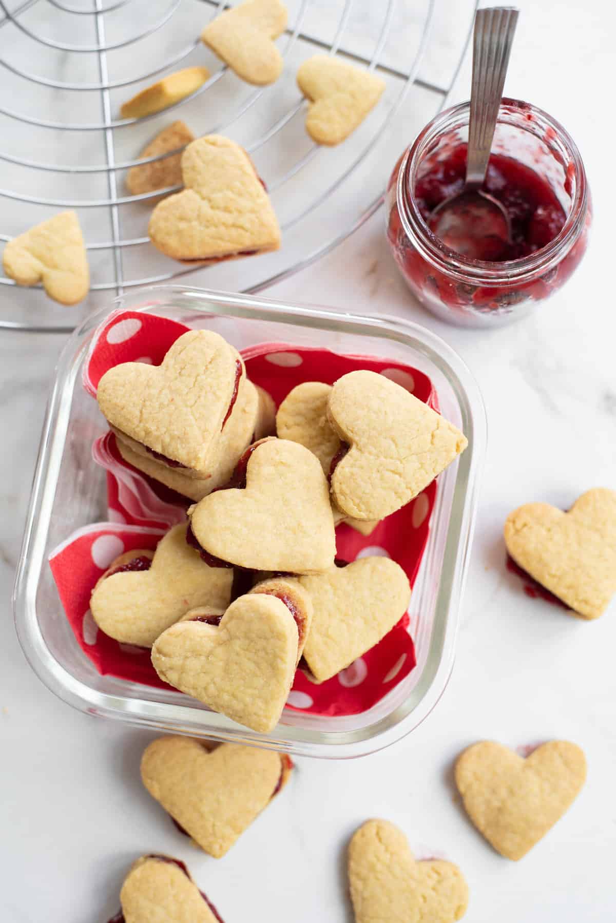 top down image of valentine's day cookies packed in a themed glass container with a side jar of strawberry jam. 