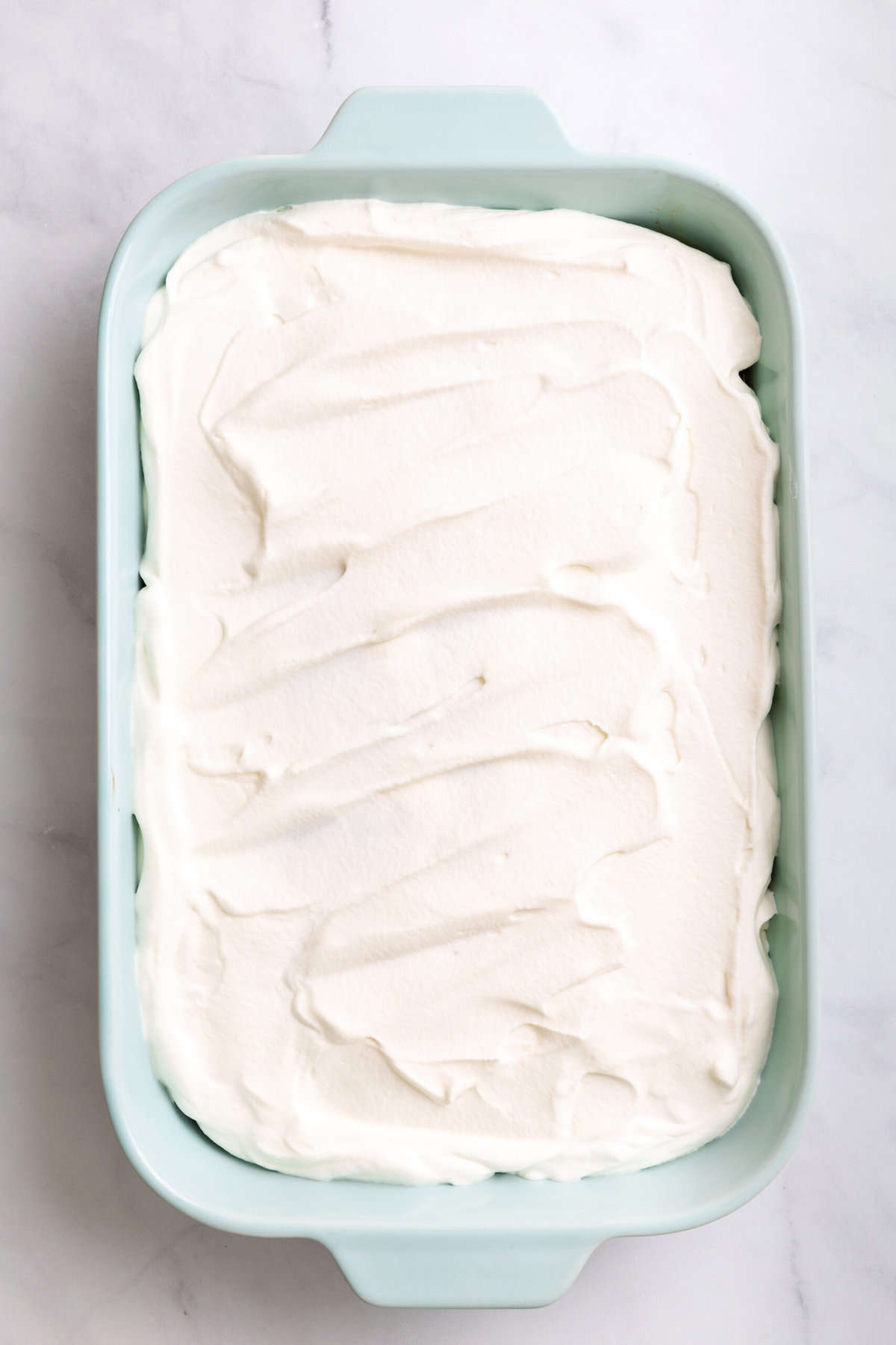 top down image of whipped cream topping on top of snickers cake sitting in a light blue 9x13 casserole dish. 