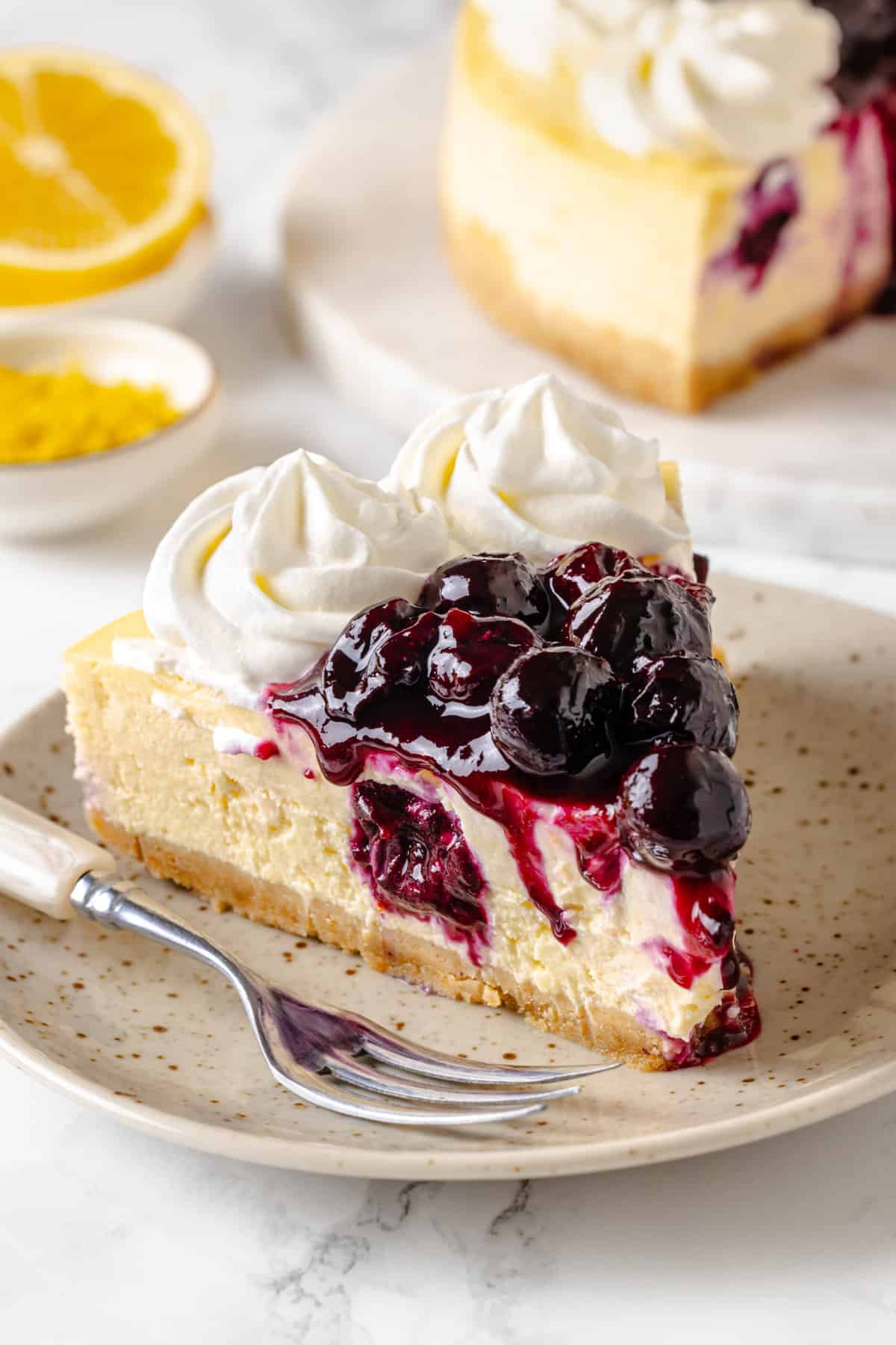 close up image of a slice of homemade lemon blueberry cheesecake sitting on a brown speckled plate. 