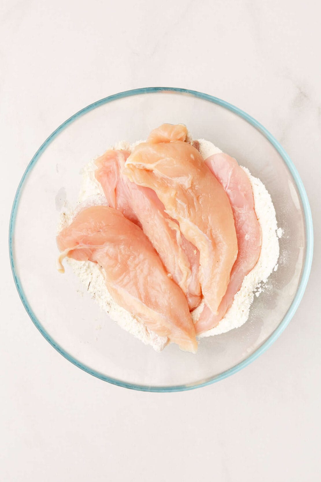 top down image of a large glass bowl with a flour mixture and raw chicken tenders. 