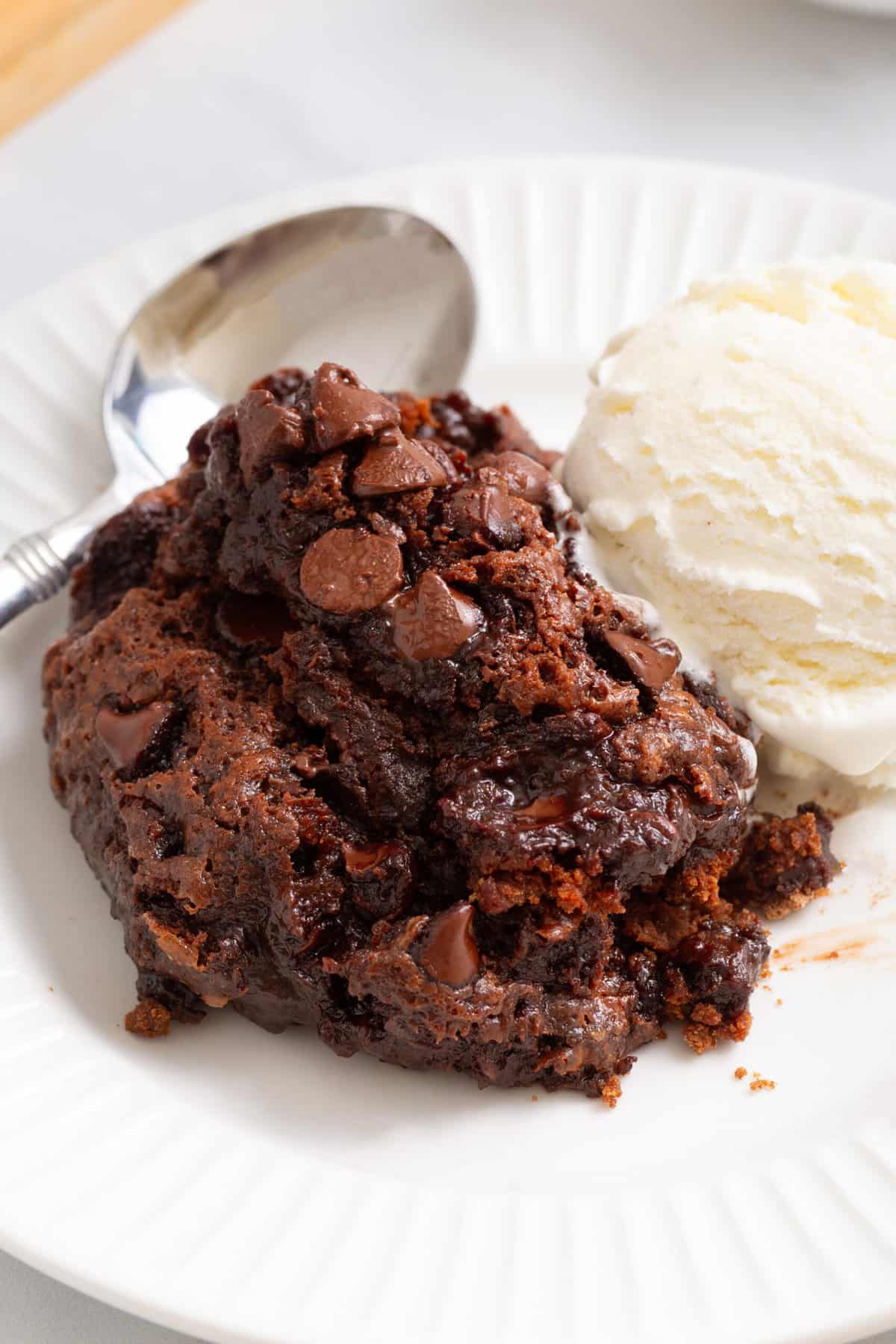 serving of chocolate dump cake served on a white round plate with a scoop of vanilla ice cream and a silver spoon. 