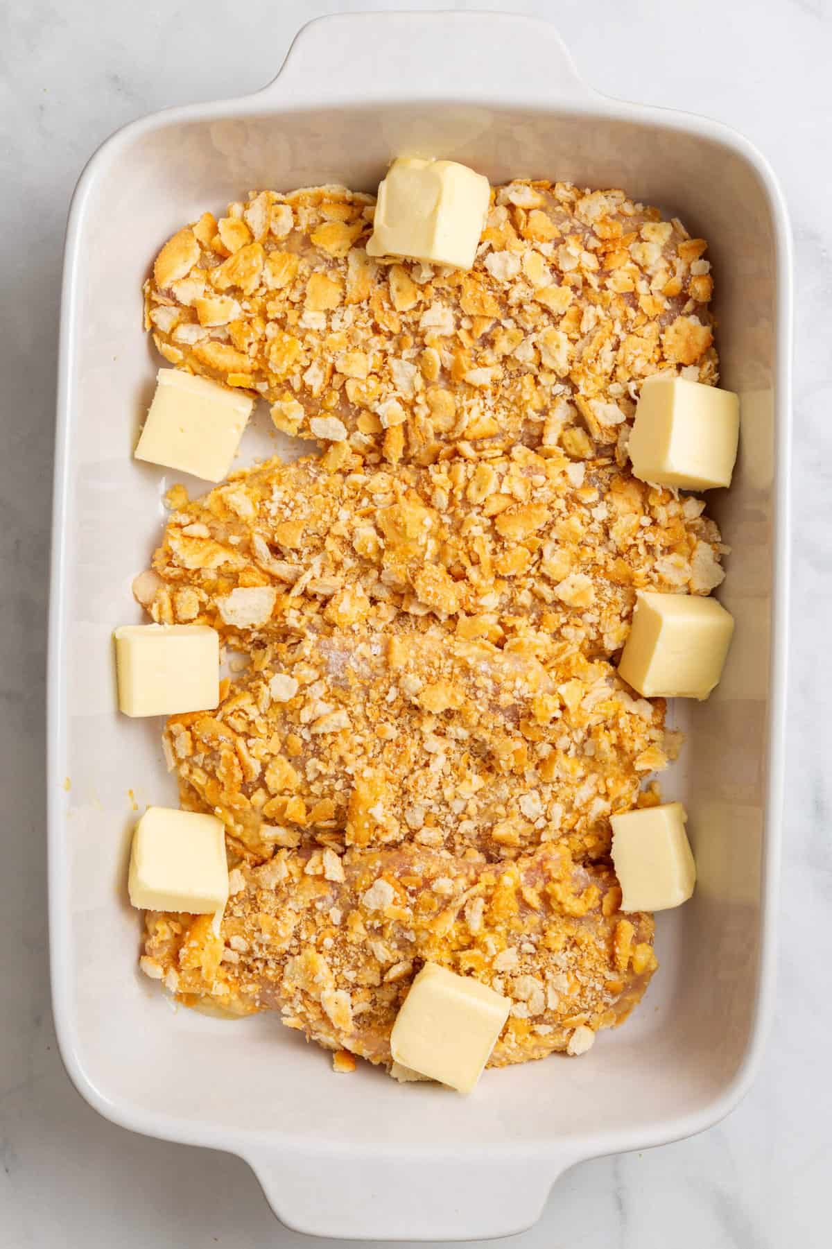 top down image of a chicken breast covered in flour mixture and egg dredge and covered in crushed ritz crackers sitting in a 9x13 casserole dish with cubes of butter.