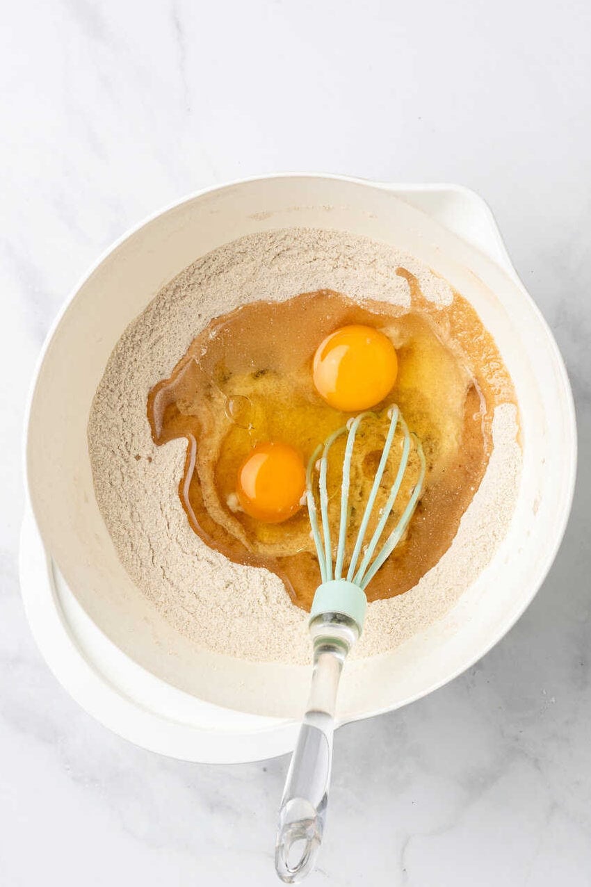 top down image of carrot cake muffin dry and wet ingredients unmixed sitting in a white bowl with a whisk. 