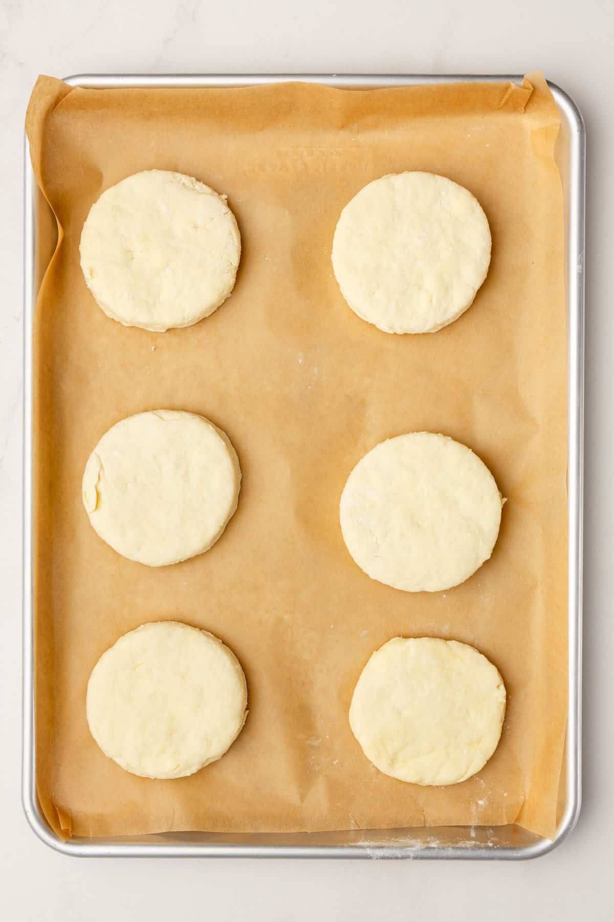 top down image of 6 circle cookie cut popeyes biscuit dough arranged on a parchment-lined baking tray. 
