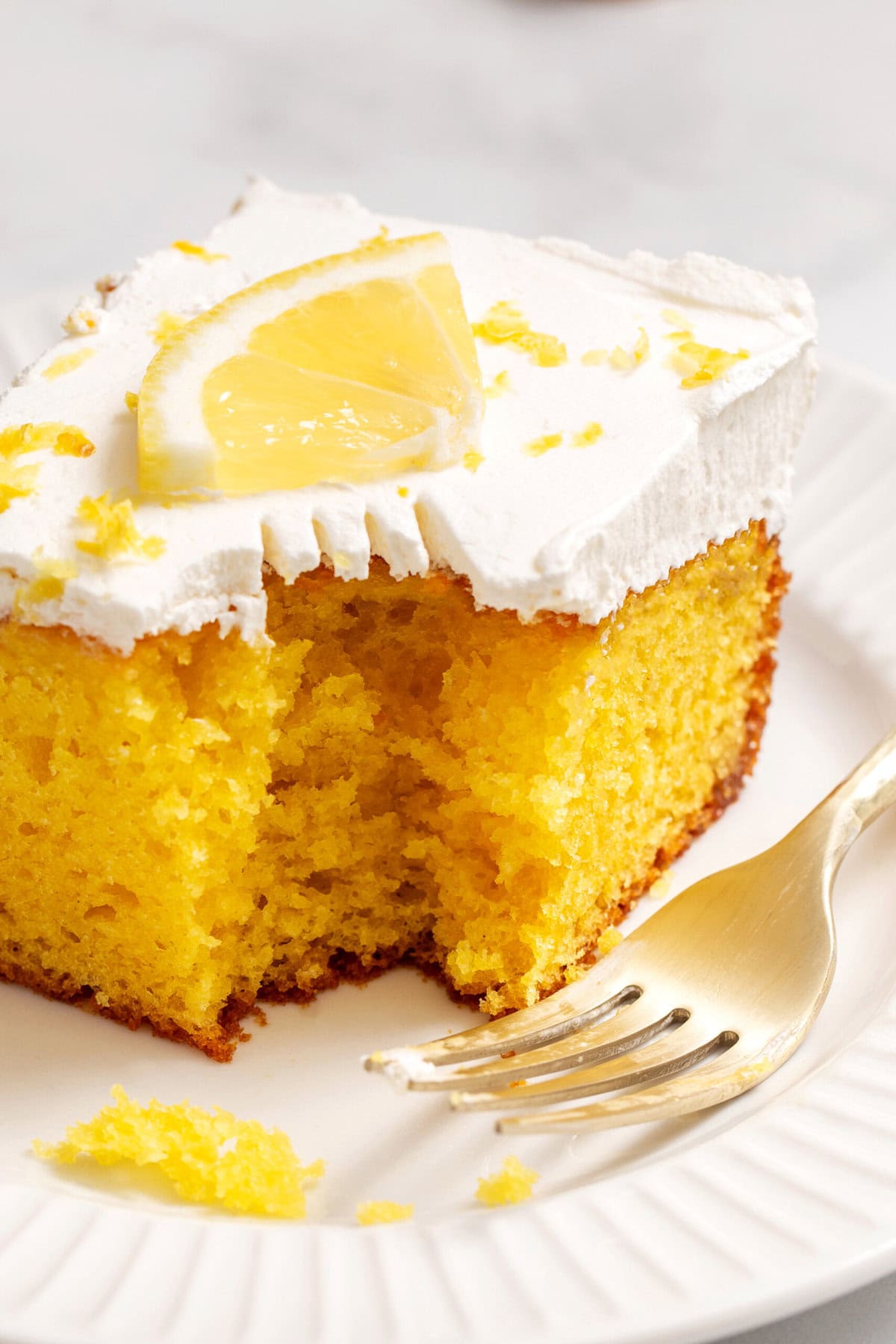 Close-up image image of a square slice of lemon poke cake, sitting on a white round plate with a bite taken out of it.