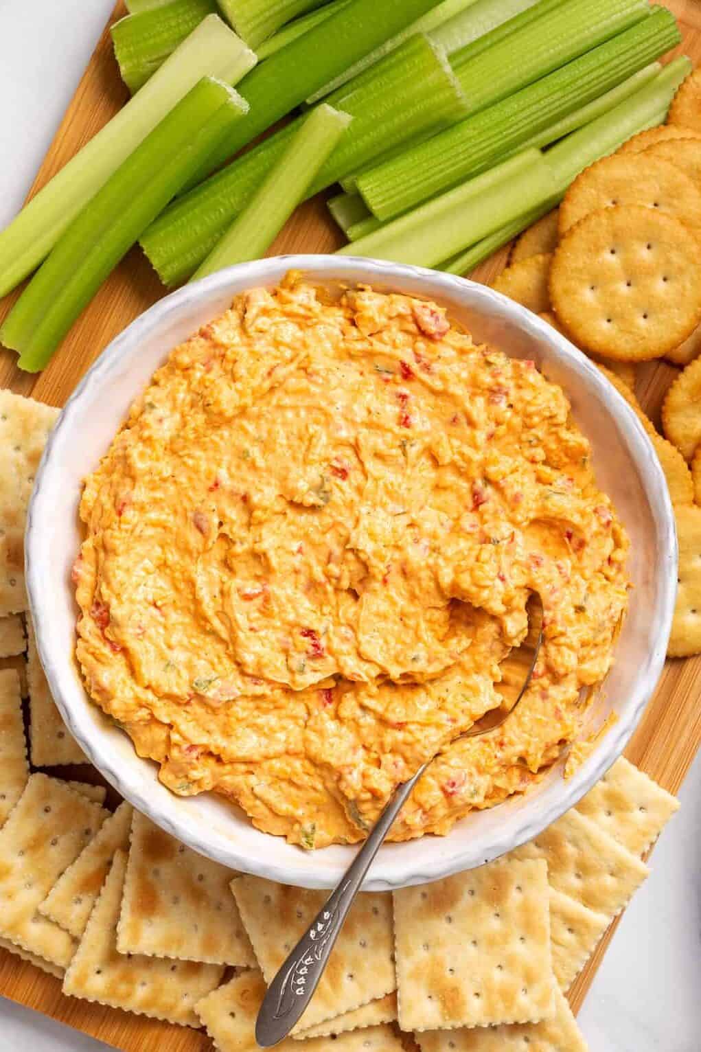 top down image of a bowl of pimento cheese dip sitting on a wooden board surrounded with ritz crackers, salted crackers, and celery sticks. 