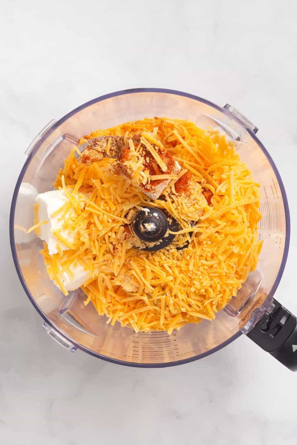 top down image of a food processor with cream cheese, shredded cheddar cheese, mayonnaise, paprika, garlic powder, onion powder, cayenne pepper.