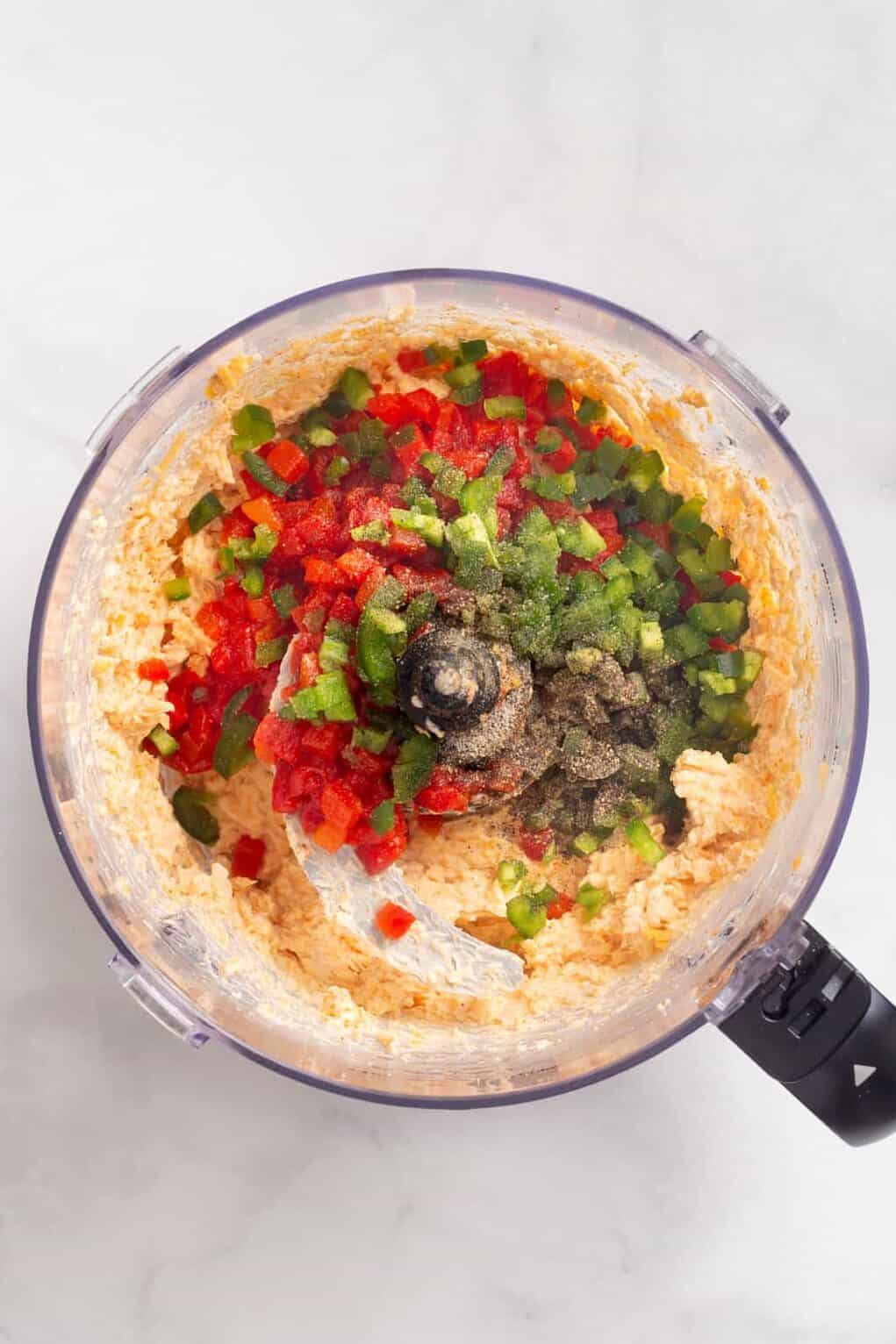 Top down image of a food processor with pimento cheese dip ingredients with the addition of pimento peppers, jalapeños, salt, and pepper.