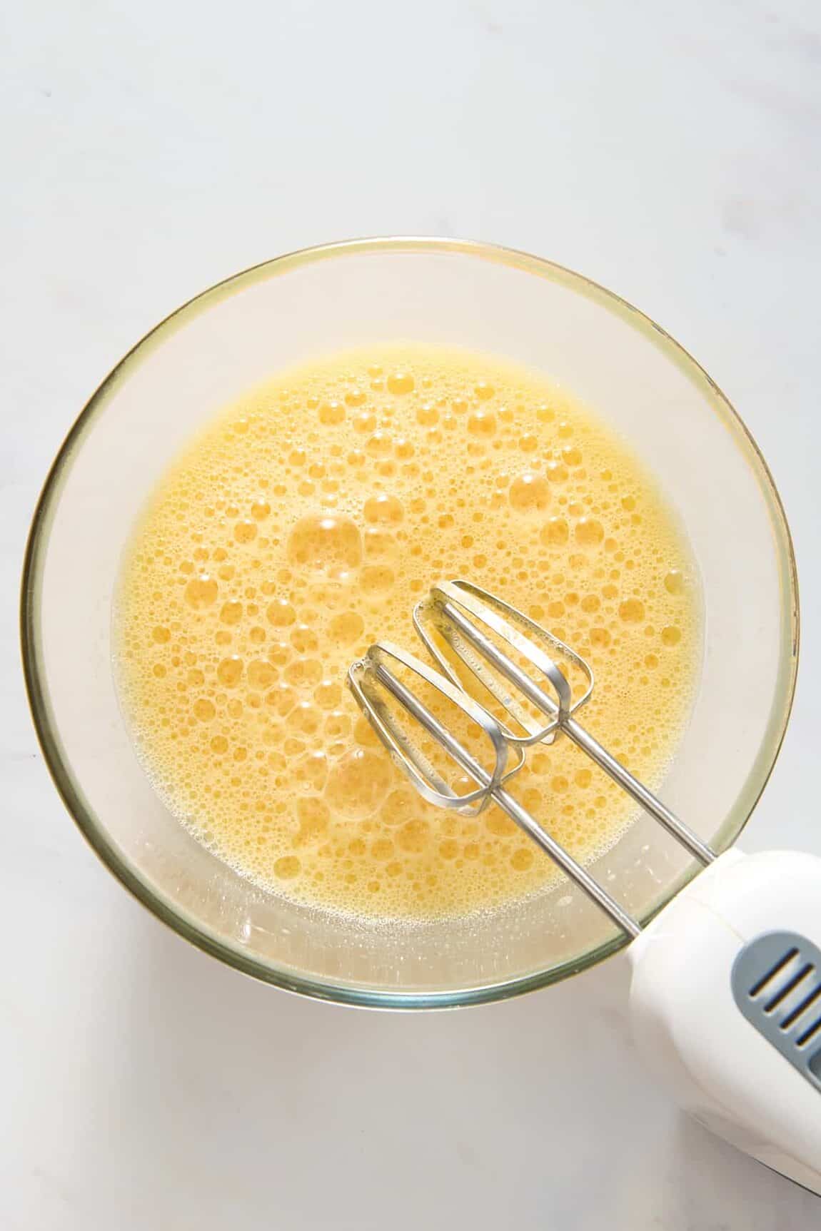 Image of an egg mixture, sitting in a large glass mixing bowl with an electric handheld whisk.