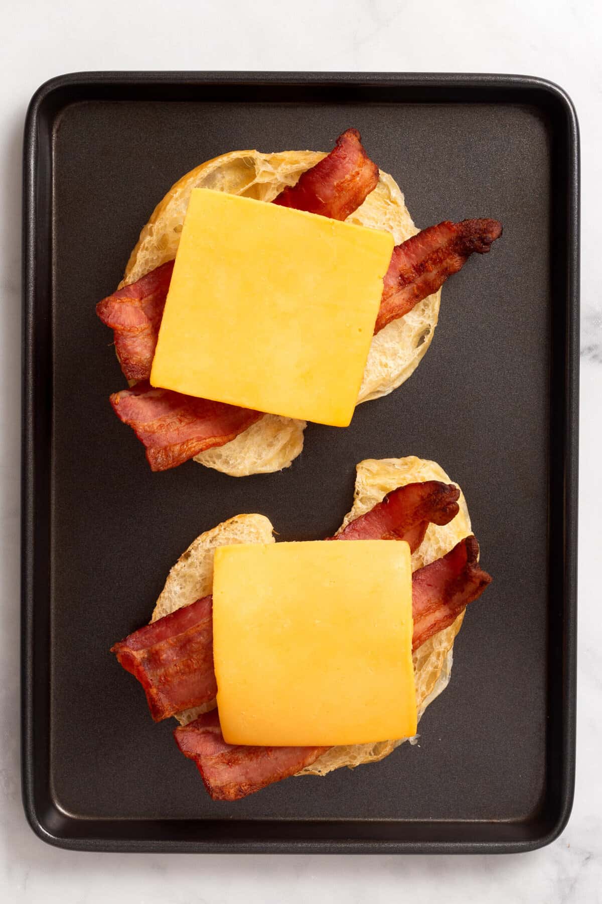 Top down image of a croissant, cut in half layered with two slices of bacon on each croissant, half and a slice of cheddar cheese on each half sitting on a baking tray.