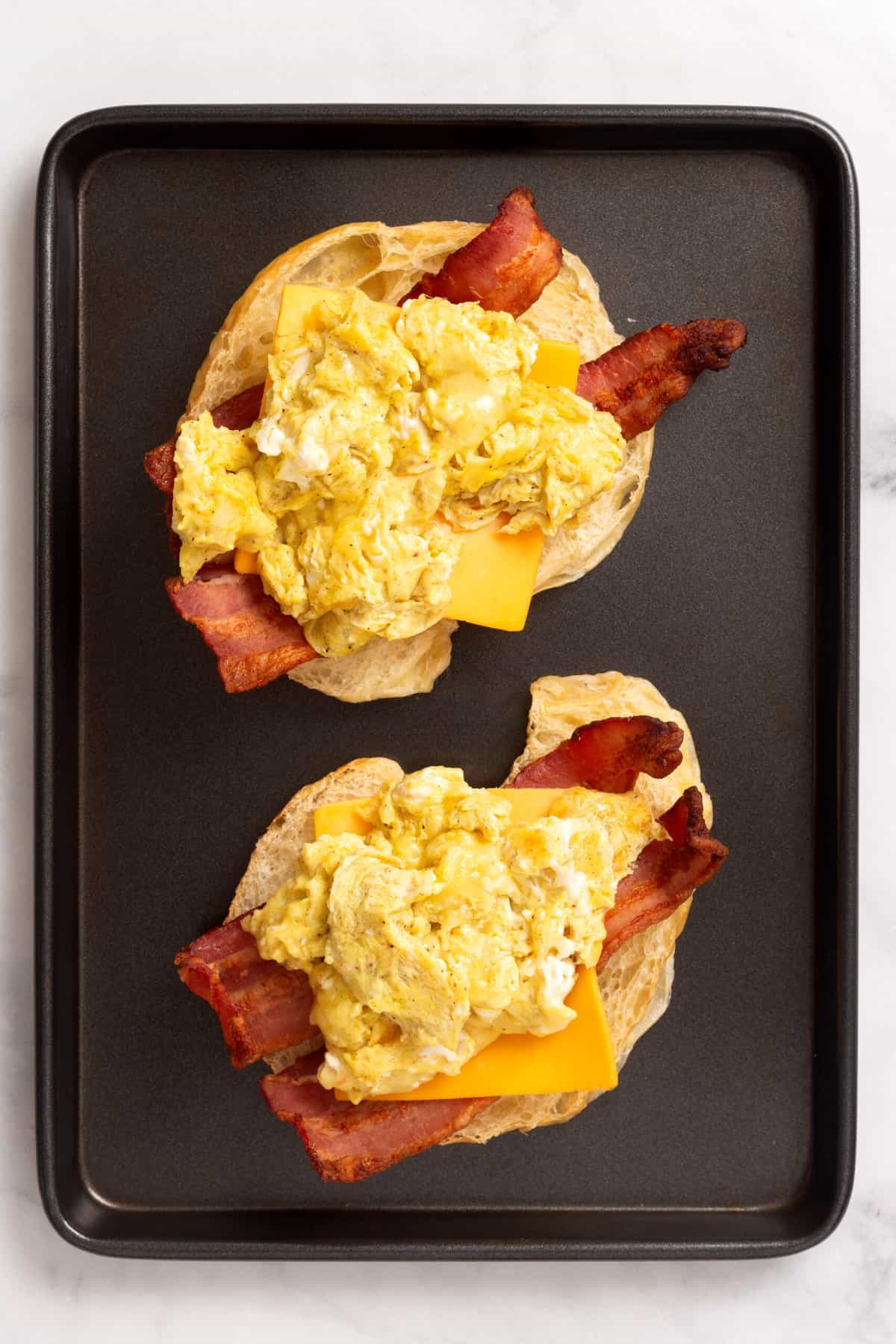 Top down image of a croissant, cut in half layered with two slices of bacon on each croissant half, a slice of cheddar cheese and scrambled, eggs on each half sitting on a baking tray.