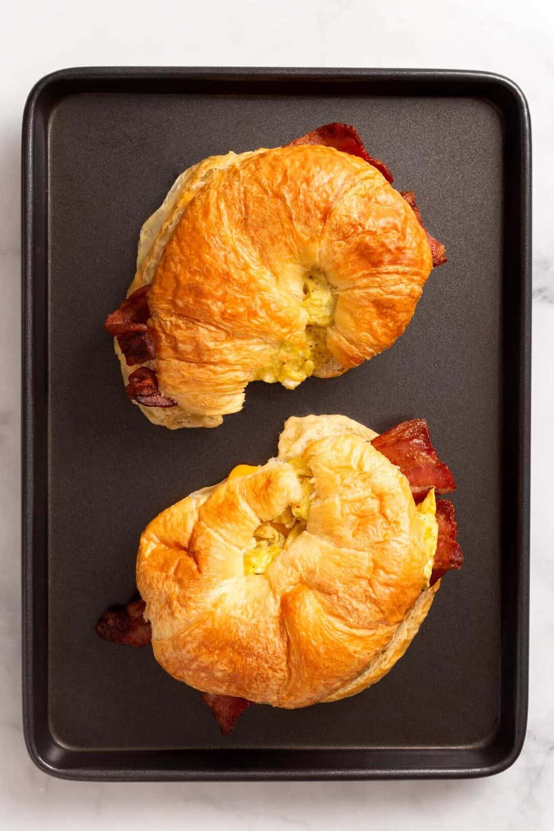 Top image of two croissant, breakfast sandwiches, sitting on a baking tray.