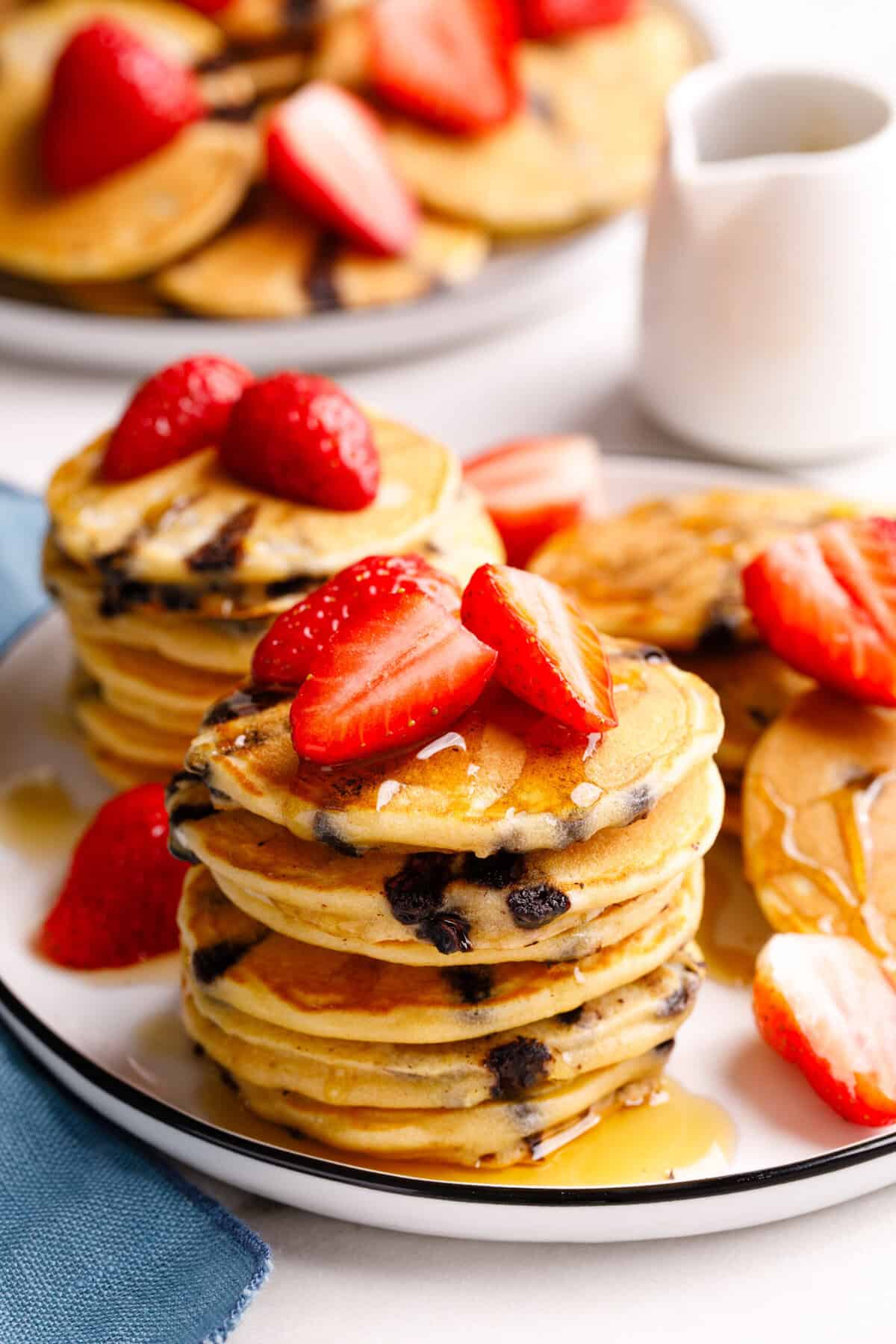 Two stacks of mini chocolate chip pancakes, sitting on a white round plate topped with fresh strawberries and syrup.