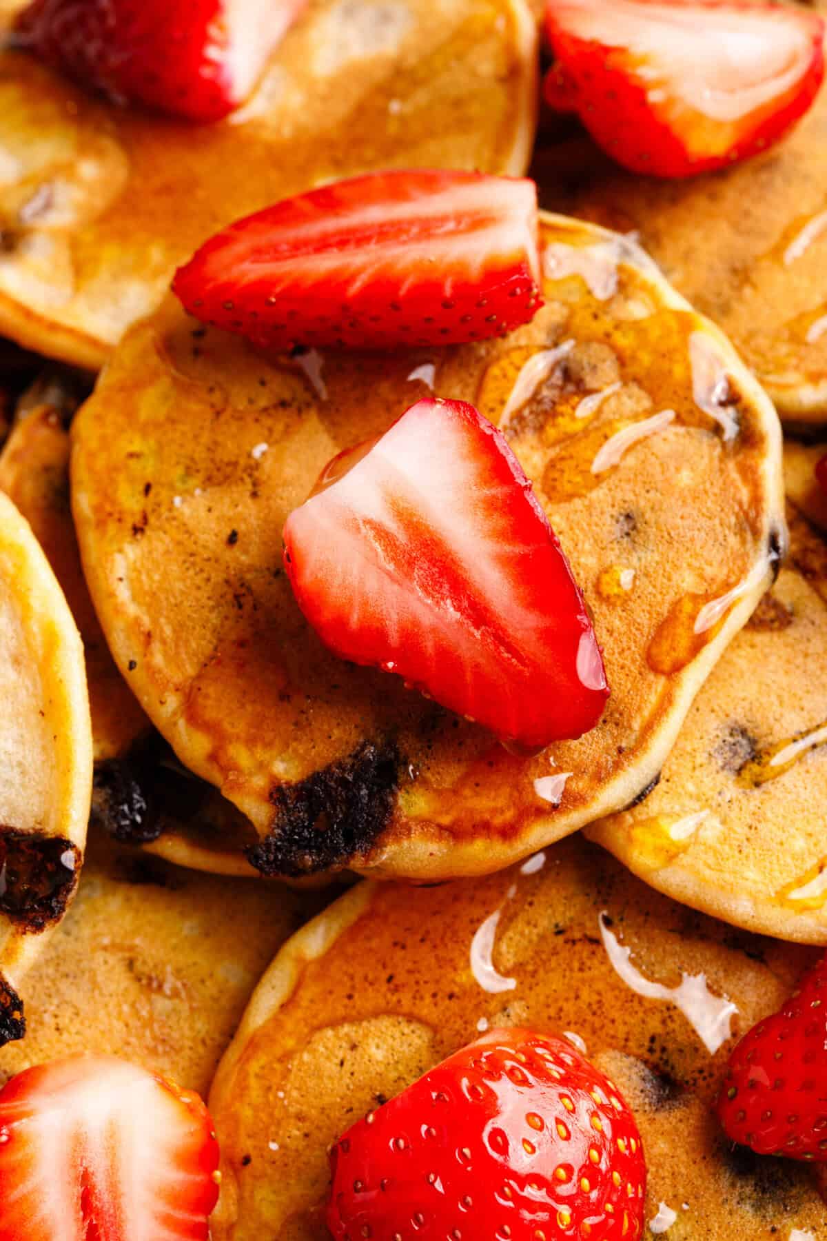 Close-up image of a plate of mini chocolate chip pancakes topped with fresh strawberries, and syrup.