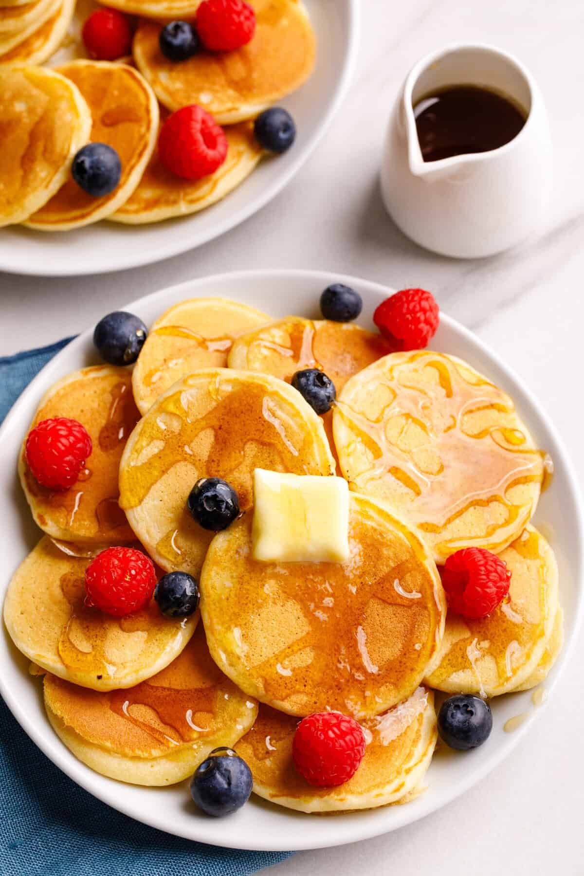 Top down image of a large round plate with silver dollar pancakes, topped with butter, syrup, and fresh berries.