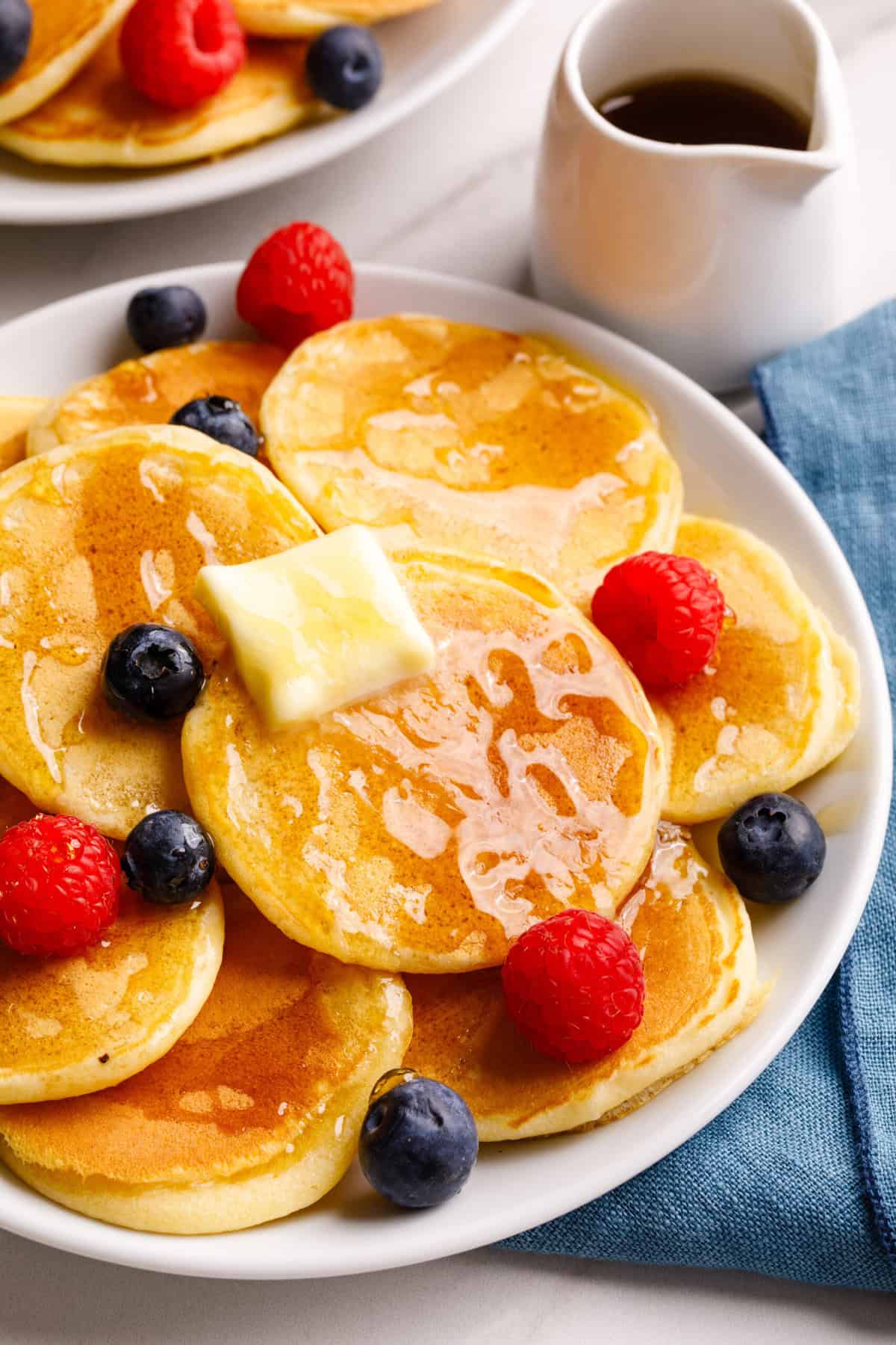 Close up of silver dollar pancakes on a white serving plate with loose berries, butter, and syrup. White jar filled with syrup and another plate in the corner with more pancakes. 