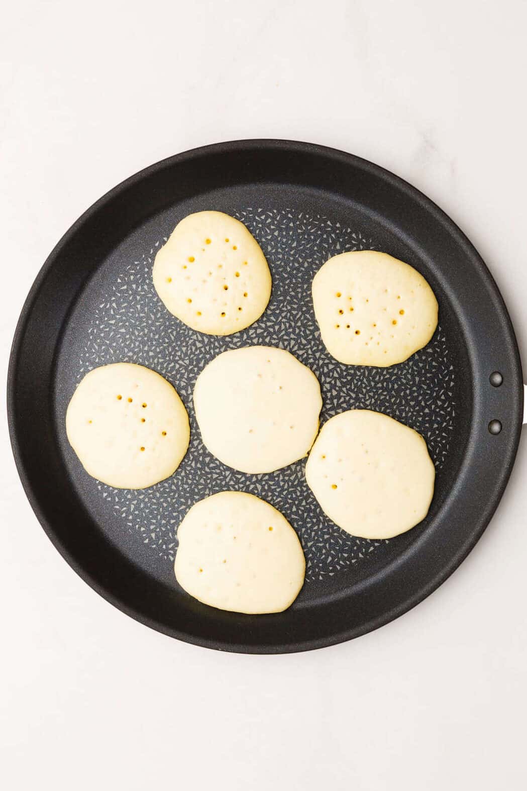 Skillet heated with melted butter and drops of silver dollar pancake batter, bubbles forming on the surface of the pancakes.