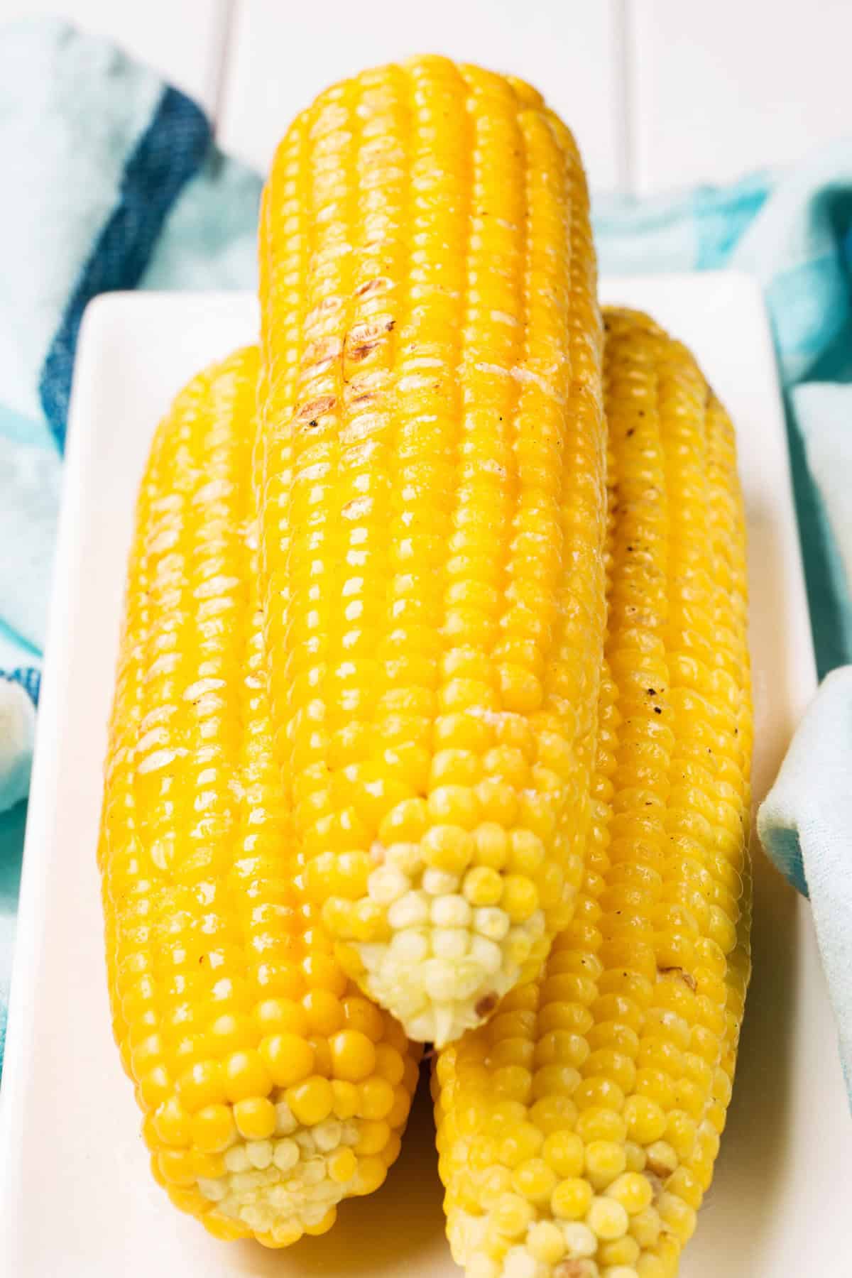 Close up of three stacked grilled corn on the cob on a white serving plate, blue and white kitchen cloth set under the plate.