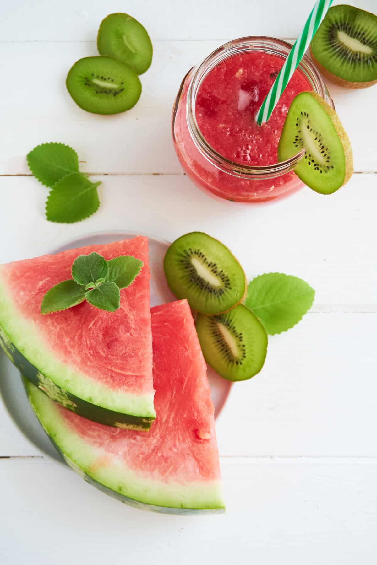 Sliced watermelon on a white side plate with sliced kiwi, overhead view of a watermelon beverage in a glass jar with a green and white striped straw and a slice of kiwi as garnish.