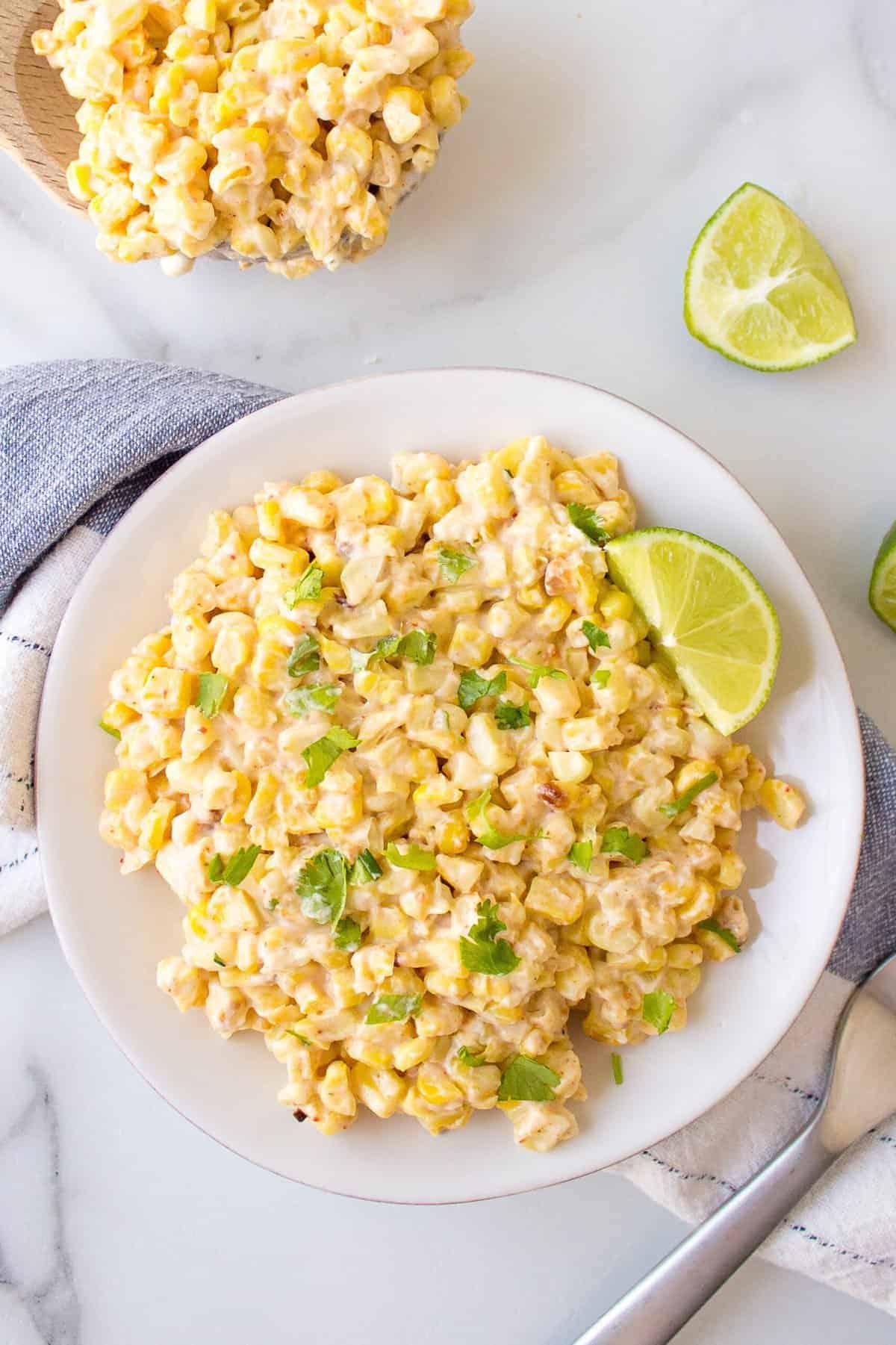 Mexican corn served on a white plate with a lime wedge, additional Mexican corn on a wooden serving spoon, a blue and white kitchen cloth underneath the plate with a metal fork off to the side.