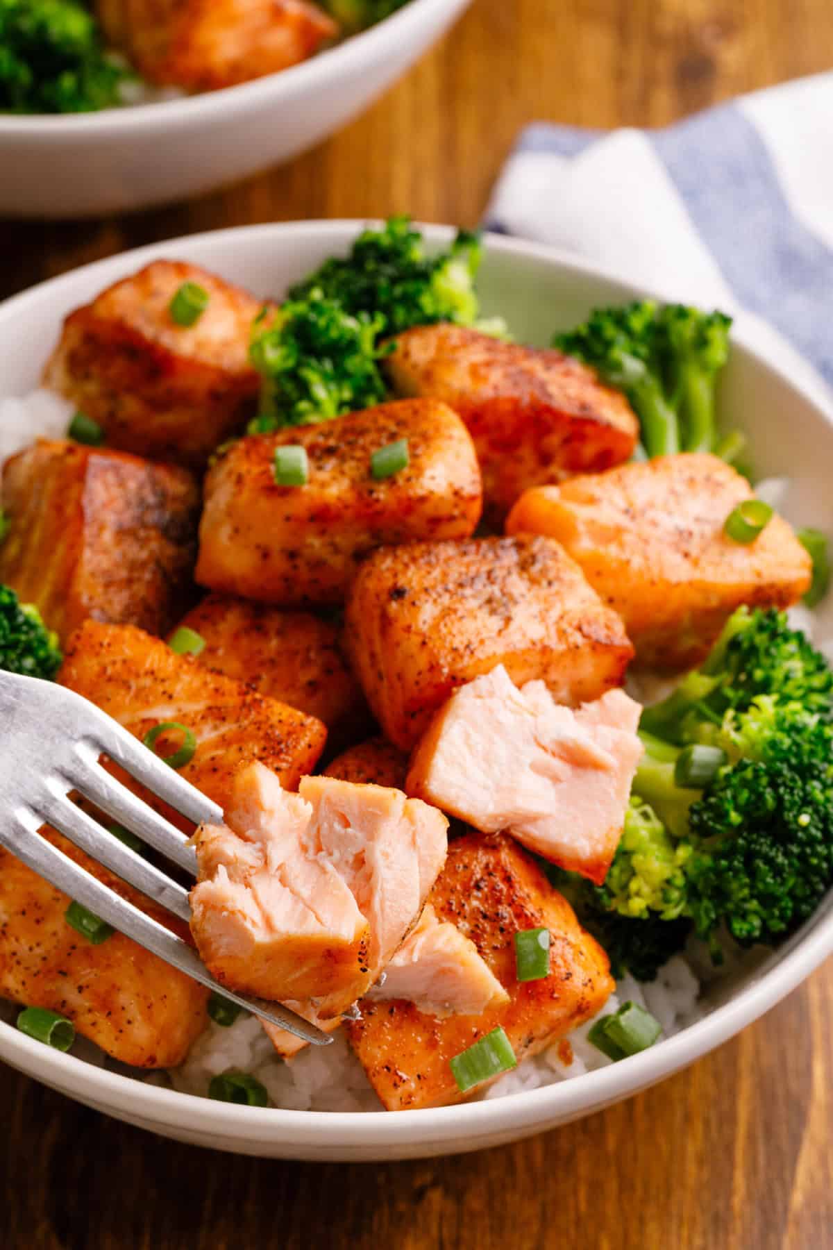 Golden salmon bites in a bowl with rice and broccoli, garnished with finely chopped green onions, silver fork piercing a salmon cube and exposing the pink flesh.
