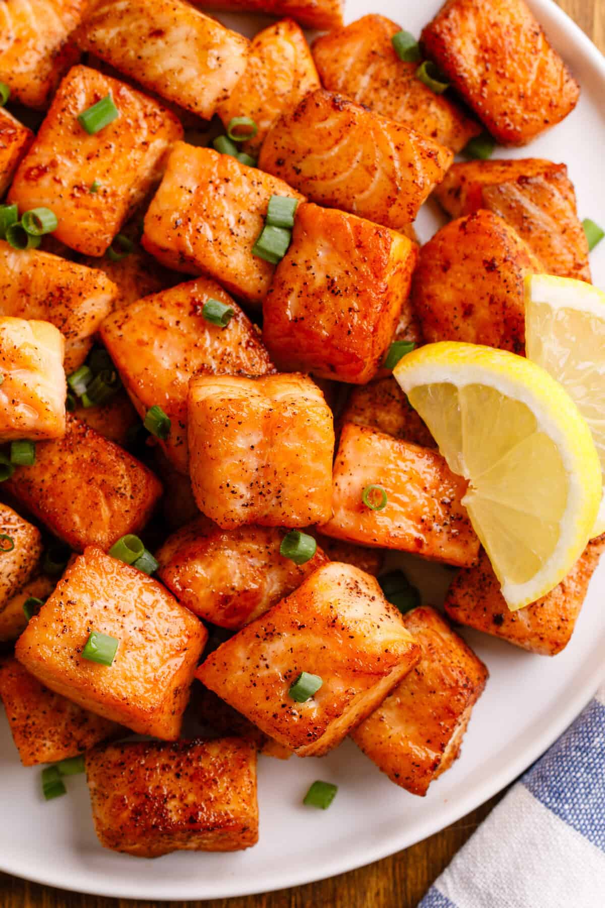 Close up of salmon bites on a white serving plate with finely chopped green onions and freshly sliced lemon.
