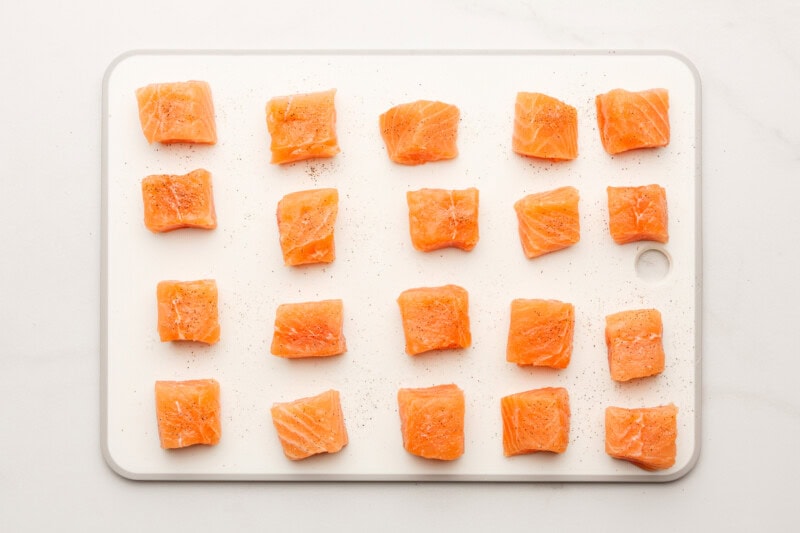 Salmon fillet cubes seasoned with salt and pepper on a cutting board.