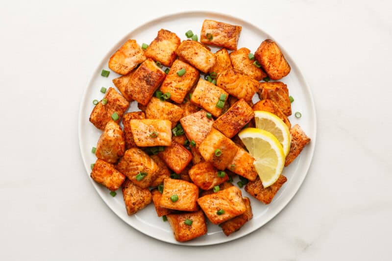 White serving plate with cooked salmon bites, garnished with freshly chopped green onions and served with lemon slices.