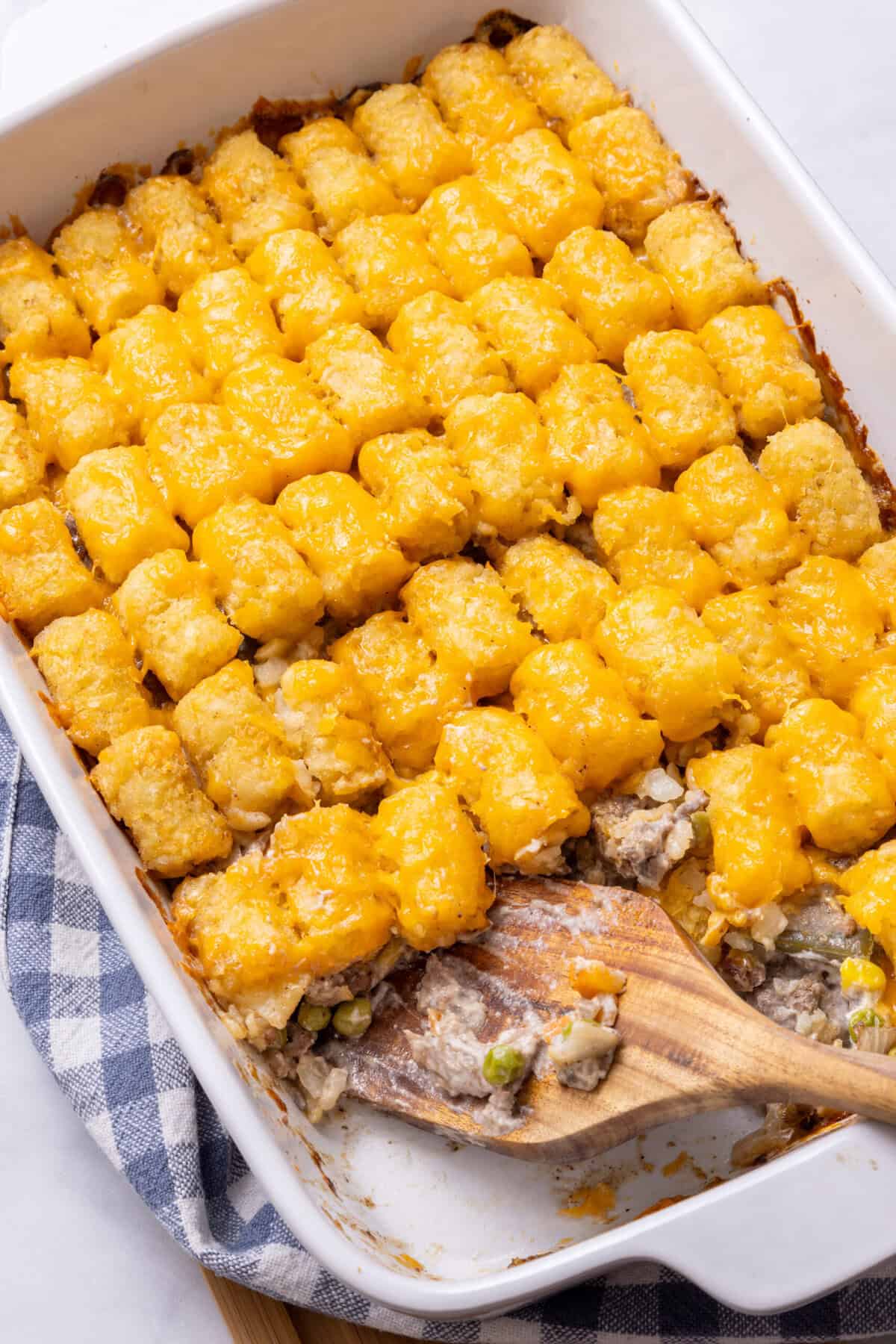 Tater Tot Casserole in a white baking dish being scooped out by a wooden spoon. 
