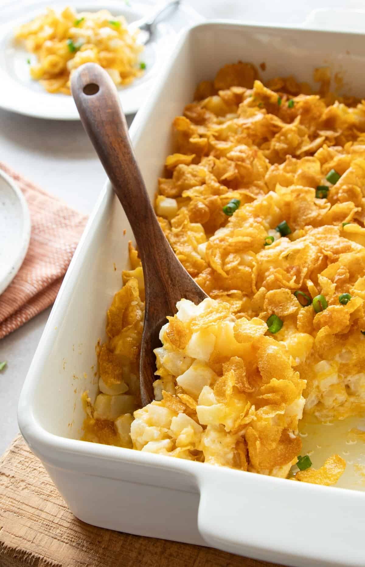 cheesy potatoes in casserole dish being scooped with wooden spoon.