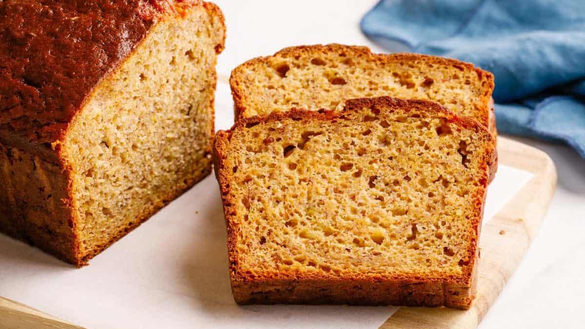Sliced 4-ingredient banana bread with the rest of the baked loaf on a wooden cutting board, blue kitchen cloth in the background.