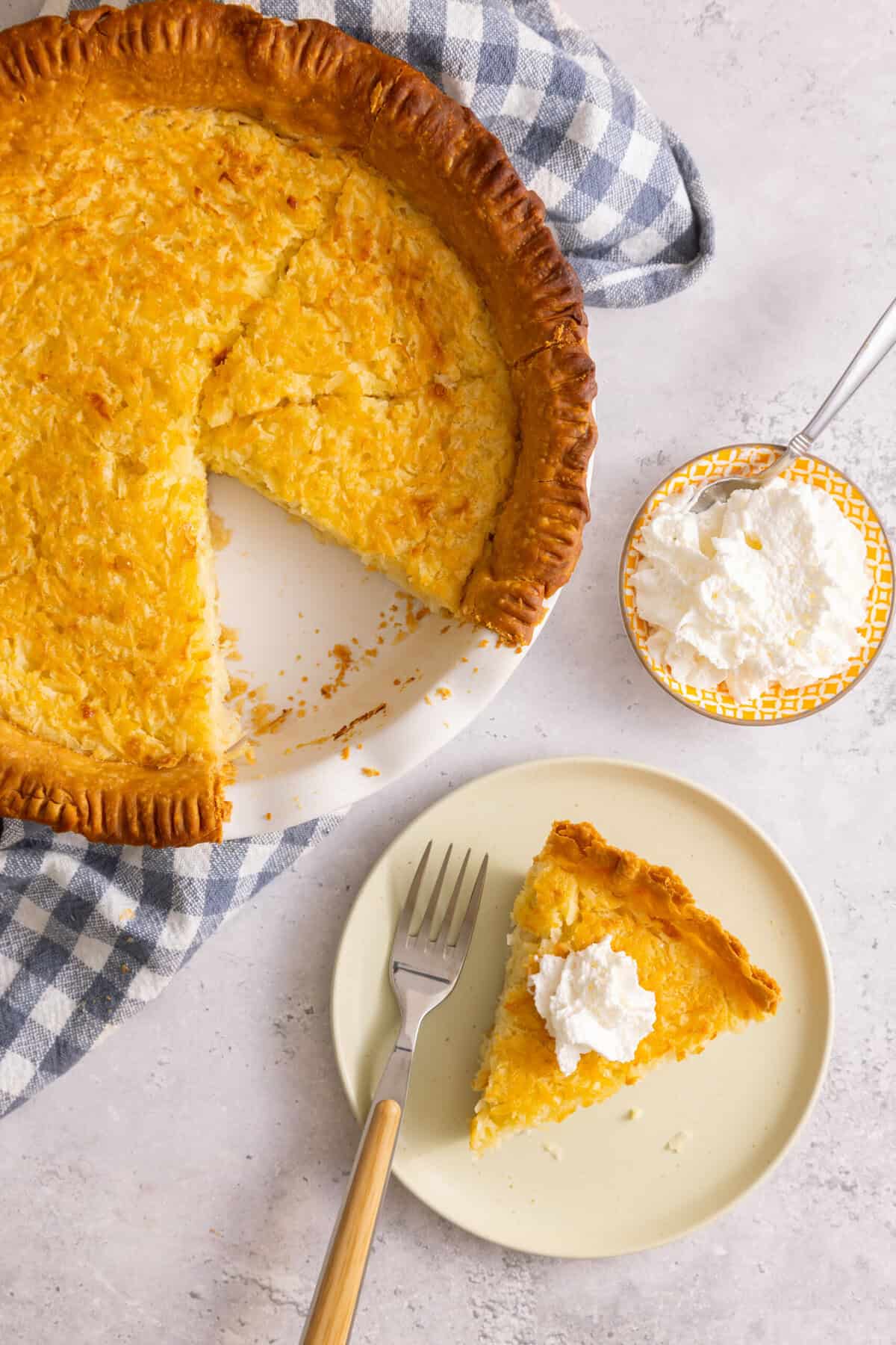 Slice of old-fashioned coconut custard pie on plate set next to pie in pie dish and bowl of whipped cream.