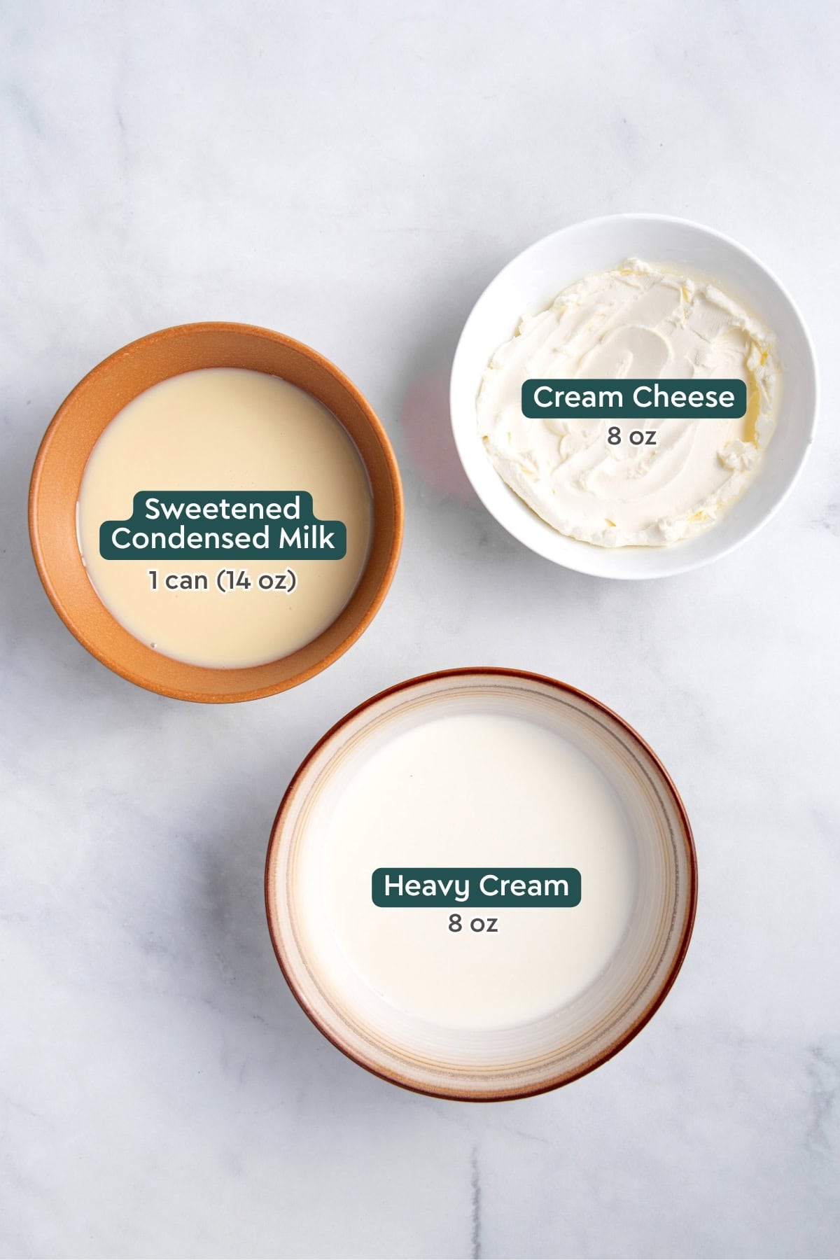 3 ingredients for no bake cheesecake are in bowls on a white countertop including softened cream cheese, heavy cream and sweetened condensed milk.