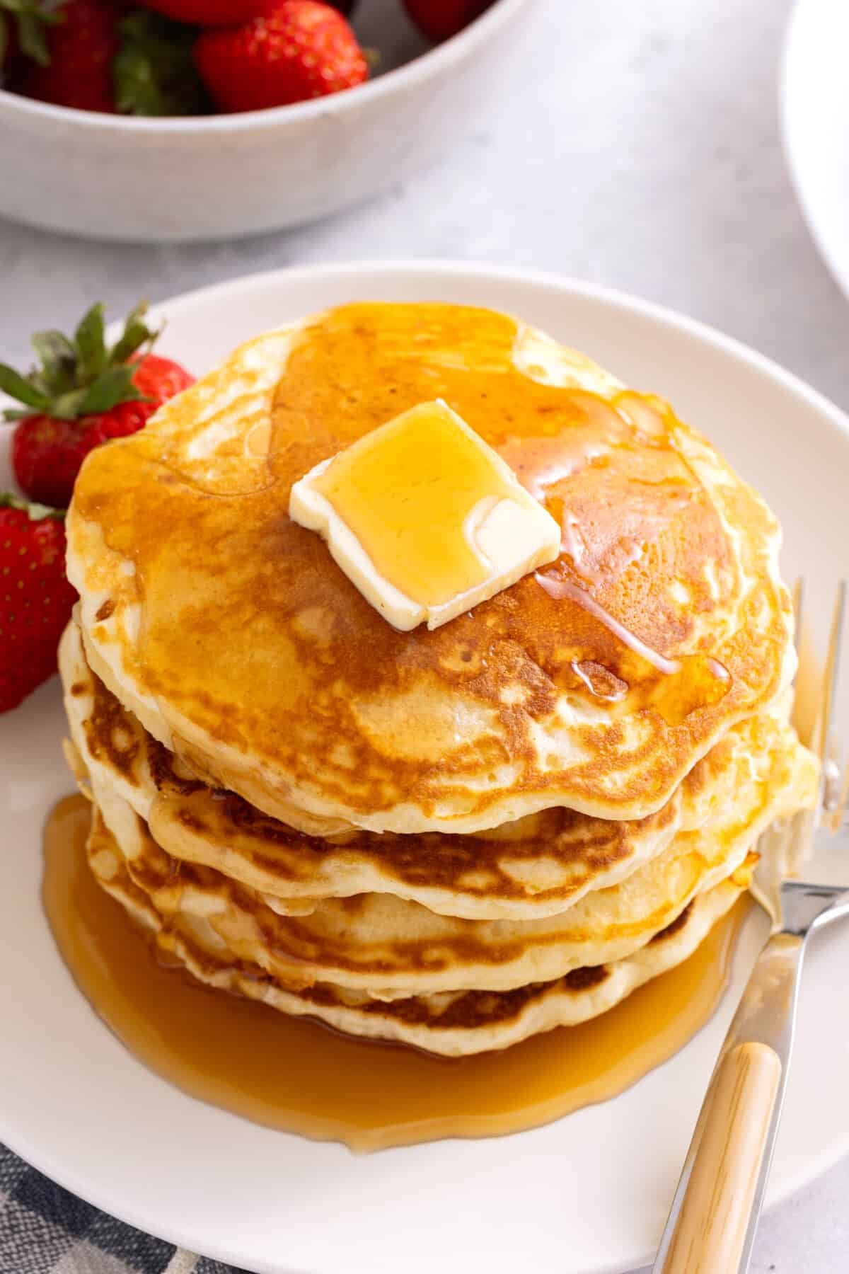Stack of fluffy homemade pancakes on plate with maple syrup and pat of butter.