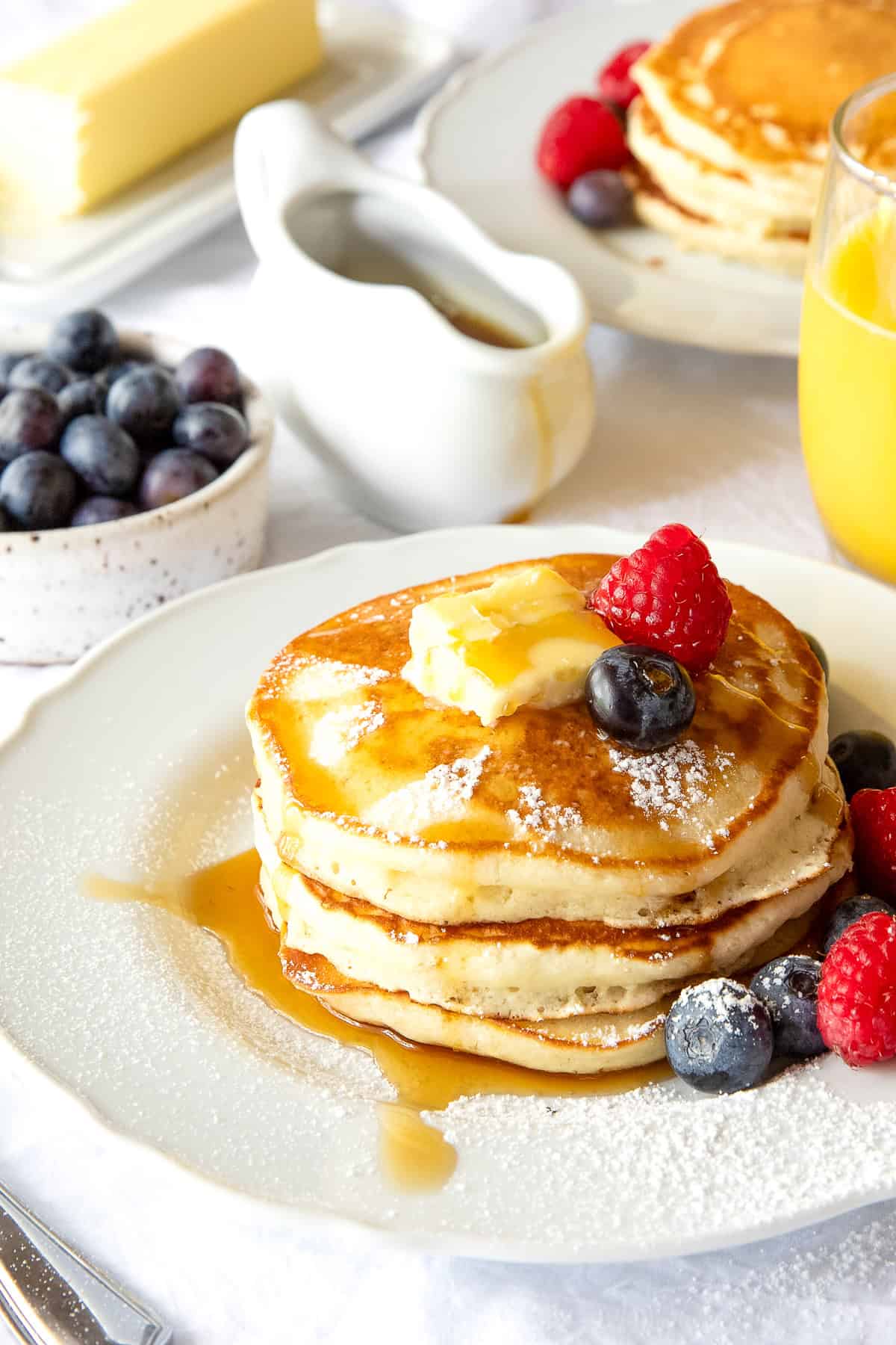 Stack of fluffy pancakes on a white plate with syrup, butter, and berries.