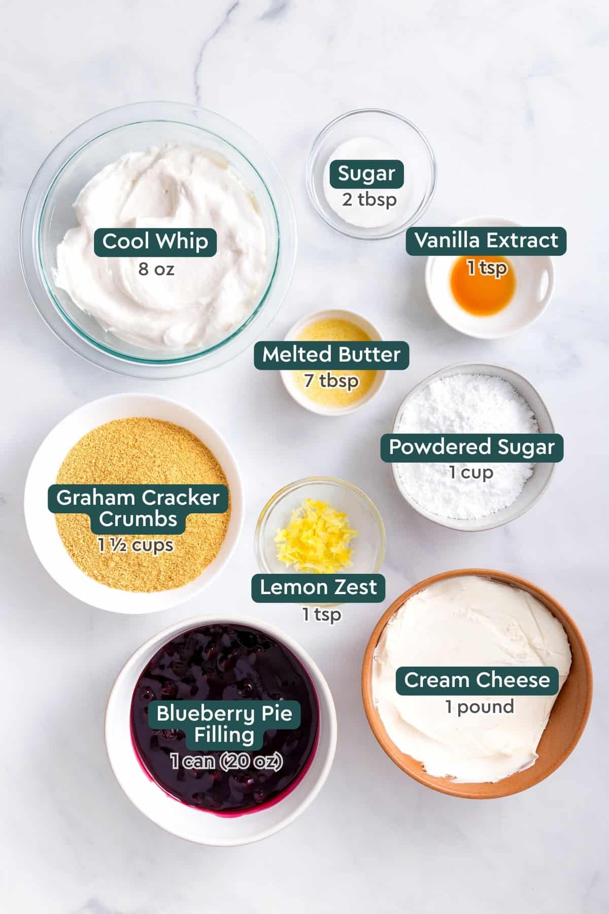 Ingredients for no bake blueberry cheesecake, including cool whip, sugar, vanilla, butter, powdered sugar, lemon zest, graham crumbs, cream cheese, blueberry pie filling.