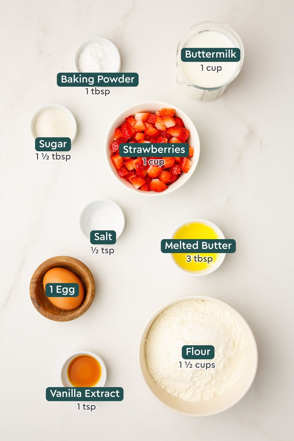 All of the ingredients for strawberry pancakes in bowls on a white countertop.