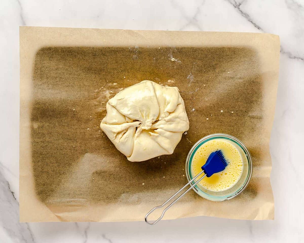 Puff pastry wrapped brie set next to bowl of egg wash with silicone brush.